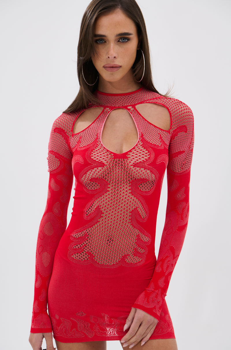 A woman with long brown hair and hoop earrings wears the HOT DAMN SEAMLESS MINI DRESS, a bright red bodycon mini with sheer mesh panels and keyhole chest cutouts. She stands before a white background, looking at the camera.