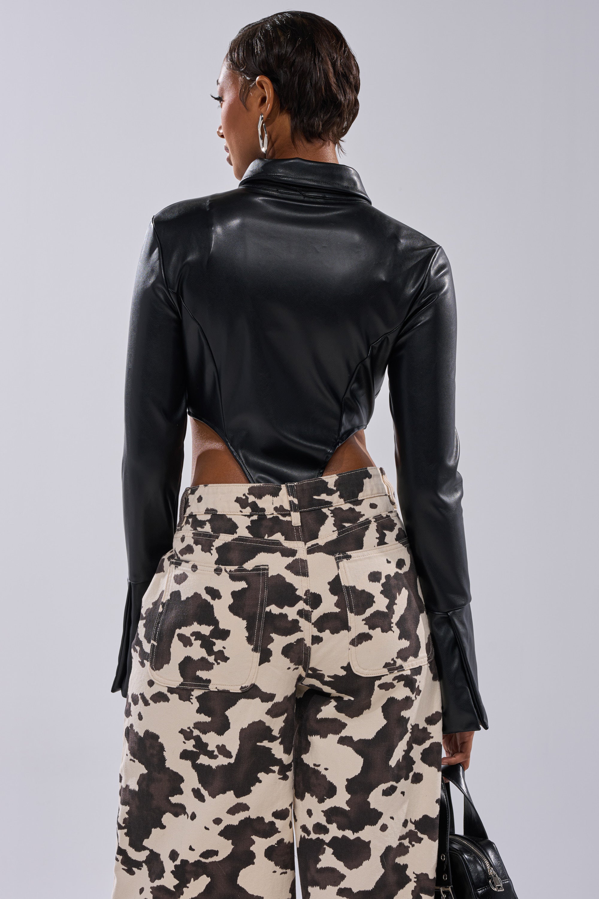 A person stands with their back to the camera, wearing the ALWAYS EXTRA BUTTON DOWN BODYSUIT, paired with high-waisted, wide-leg pants in a black and white cow print pattern, and holding a small handbag.