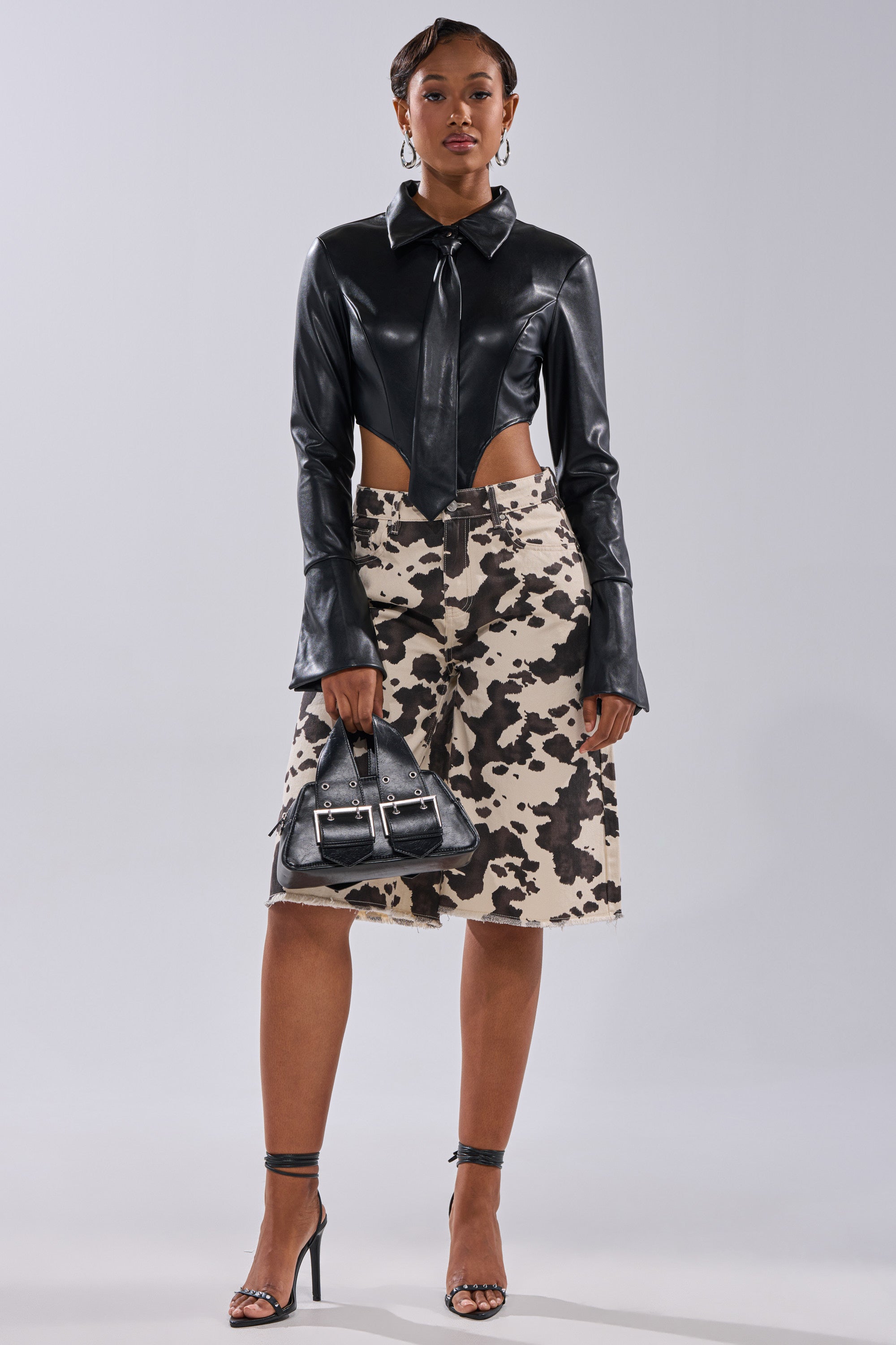 A woman stands against a plain background wearing the ALWAYS EXTRA BUTTON DOWN BODYSUIT, a curve-hugging beige and black cow-print skirt, black heels, hoop earrings, and holding a small black handbag.