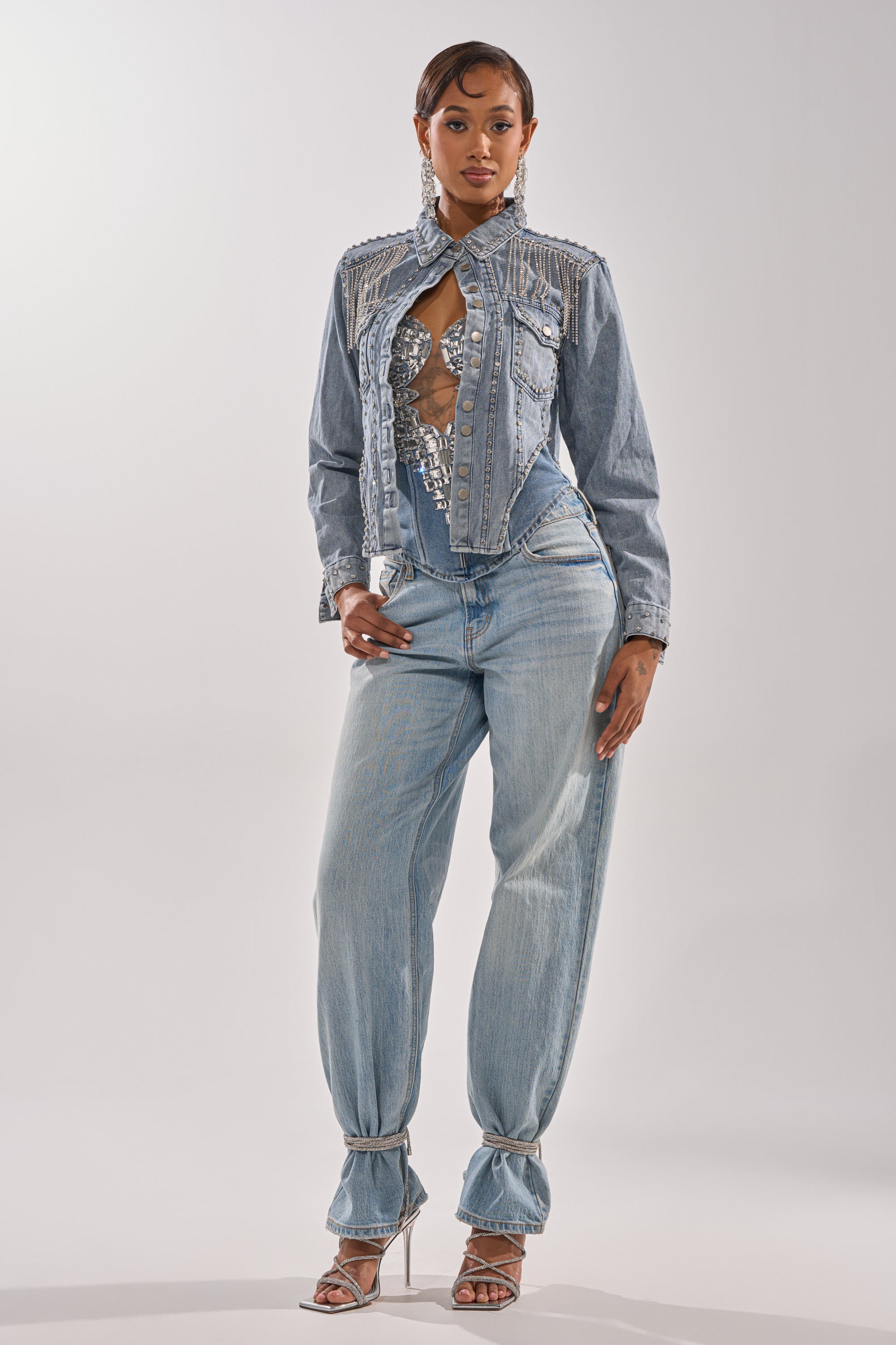 A woman poses confidently in the HOOKED ON YOU EMBELLISHED DENIM JACKET over a shimmering silver top and ankle-tied, high-waisted light blue jeans, styled with strappy heels and statement earrings. 9
