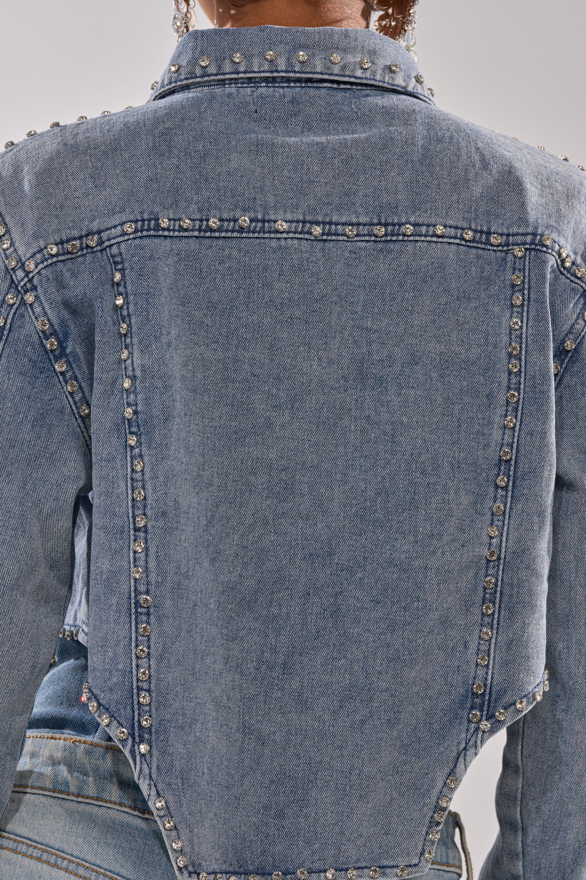 A close-up of the HOOKED ON YOU EMBELLISHED DENIM JACKET, featuring silver studs along the seams, unique angled stitching, and a faded blue wash, worn by a person with short curly hair. 11