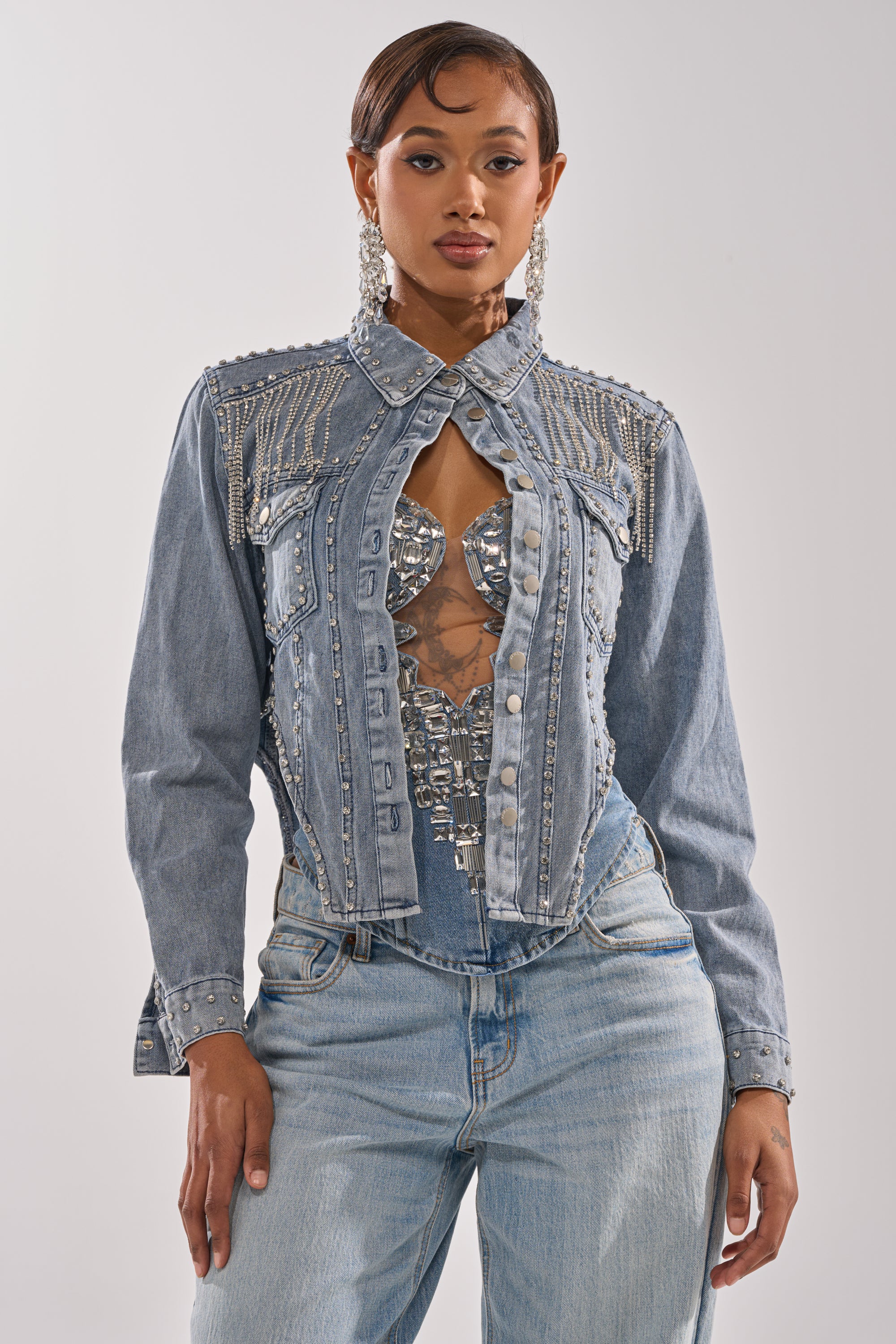 A woman poses confidently against a plain background wearing the HOOKED ON YOU EMBELLISHED DENIM JACKET with silver details and rhinestone fringe over a mirrored top, paired with light wash jeans. 1