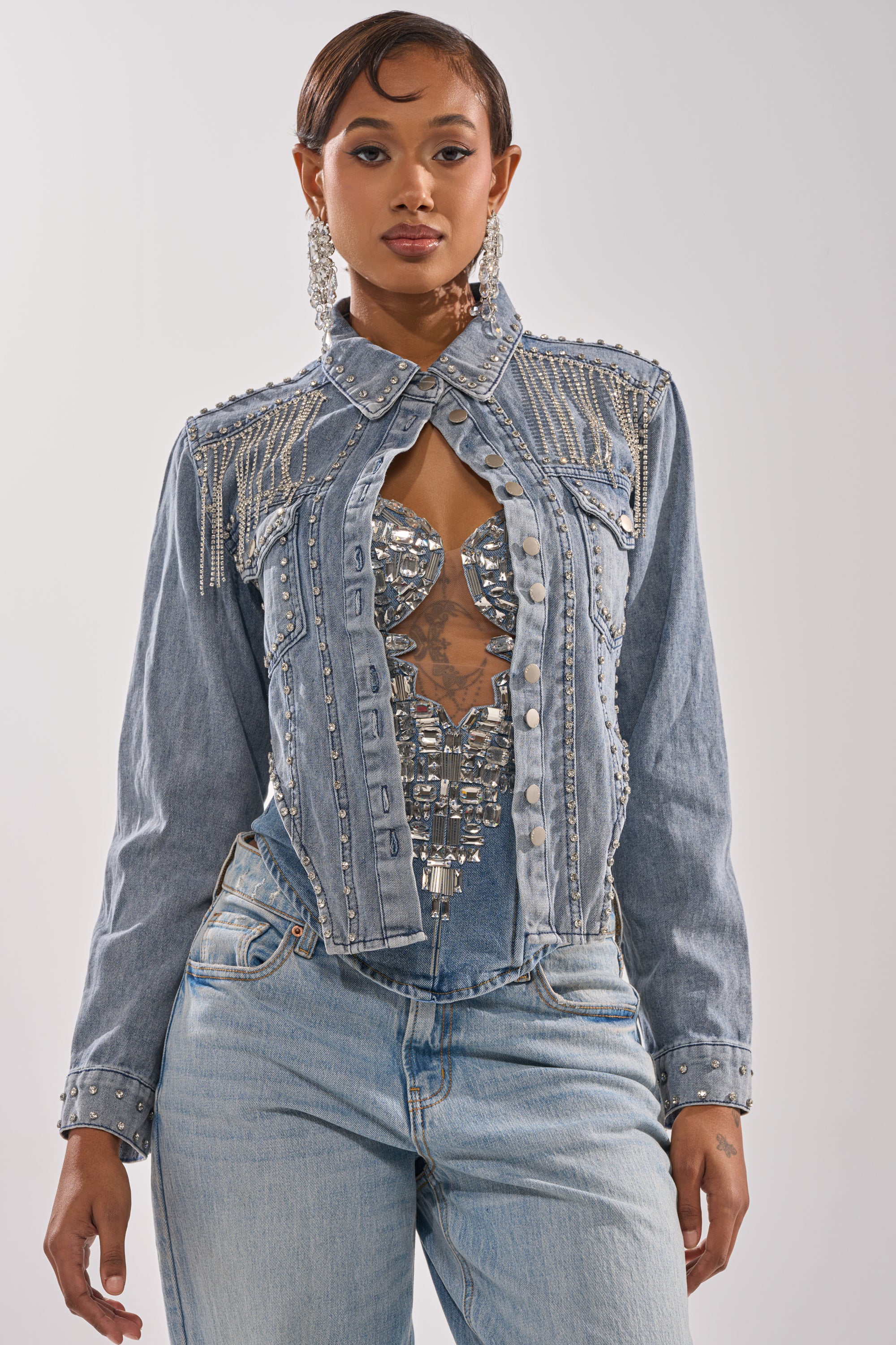 A woman poses confidently in the HOOKED ON YOU EMBELLISHED DENIM JACKET, featuring rhinestone fringe and pearl details, styled with light wash jeans and a silver mirrored corset top against a plain white background. 3
