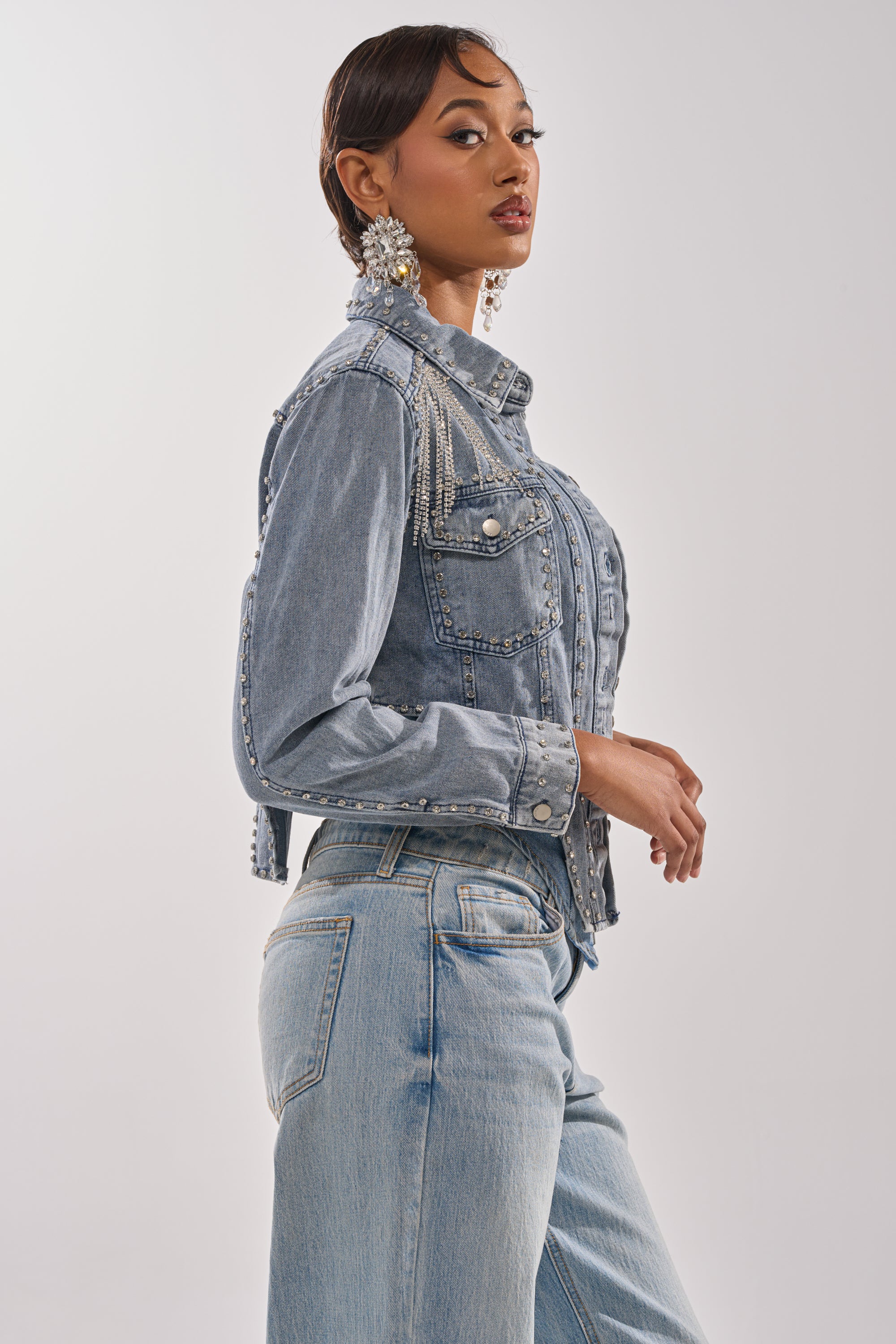 A woman with short dark hair models the HOOKED ON YOU EMBELLISHED DENIM JACKET and matching jeans, posing sideways with her head turned to the camera, accessorized with statement earrings against a plain light background. 4