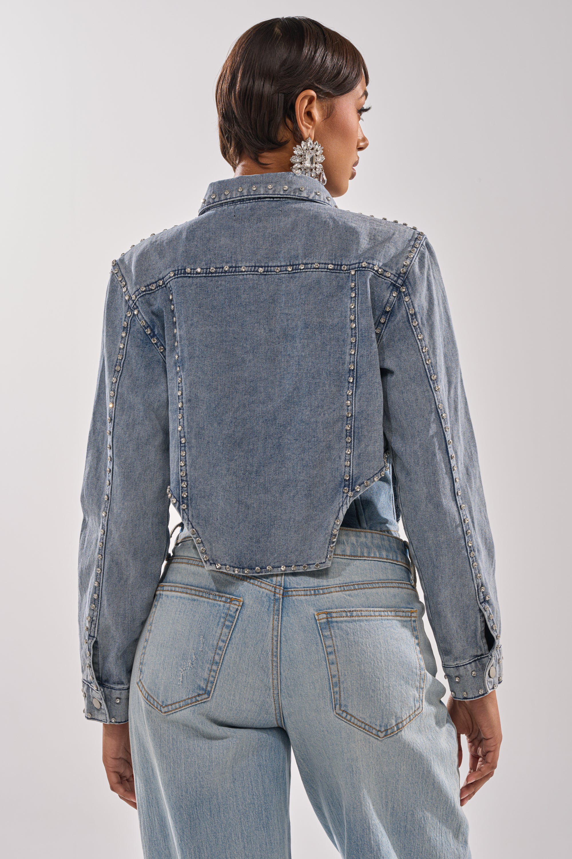 A person with short hair and large decorative earrings faces away, highlighting the HOOKED ON YOU EMBELLISHED DENIM JACKET with pearl detailing on the seams, paired with high-waisted light blue jeans. 5