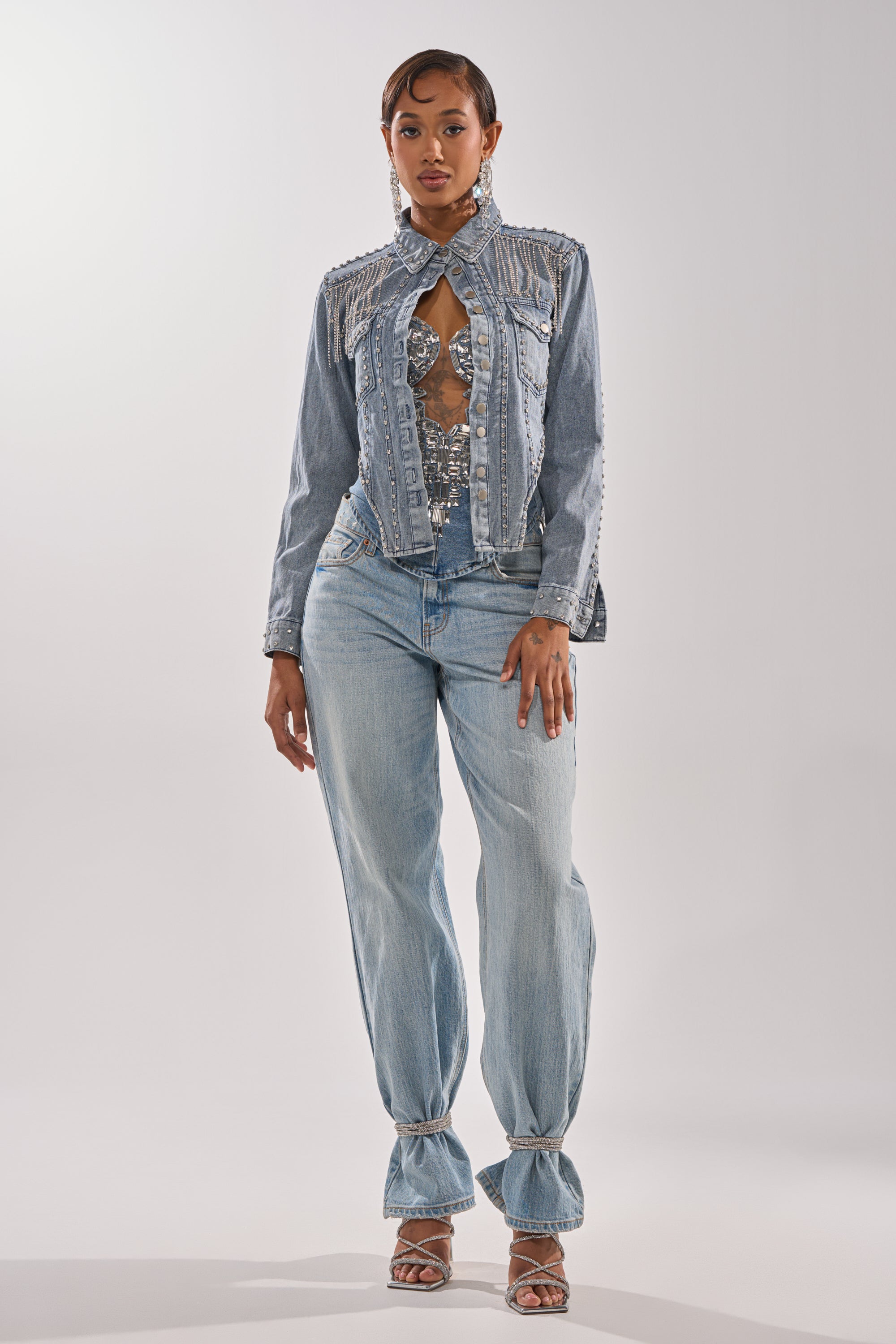 A woman stands confidently in a studio wearing the HOOKED ON YOU EMBELLISHED DENIM JACKET with decorative buttons and cutouts, matching wide-leg jeans tied at the ankles, silver heels, and statement earrings. 8
