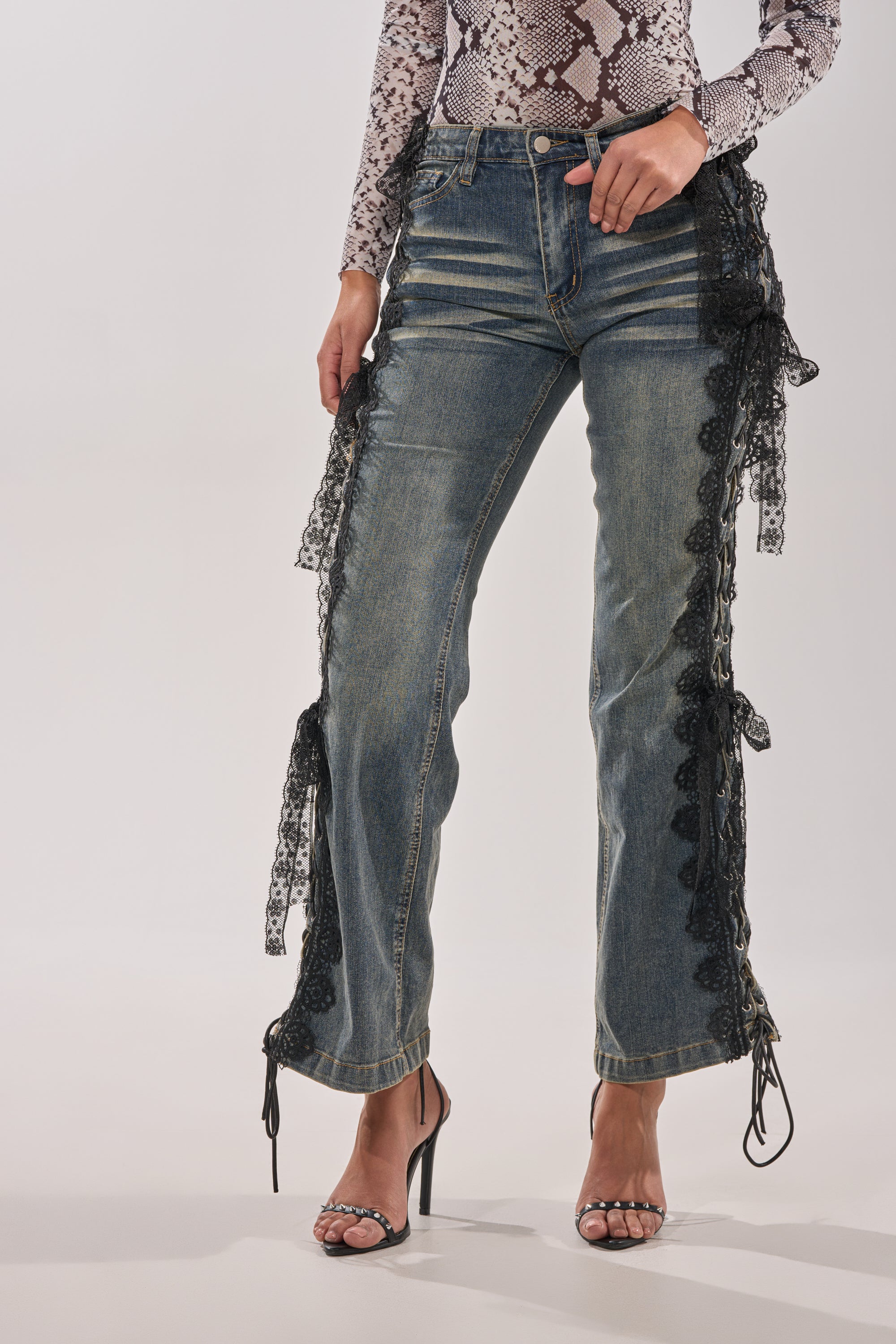 A person stands against a plain background wearing ELIJAH FLARE LACE DENIM jeans with lace-up sides, a long-sleeve snakeskin print top, and black high-heeled sandals. 0