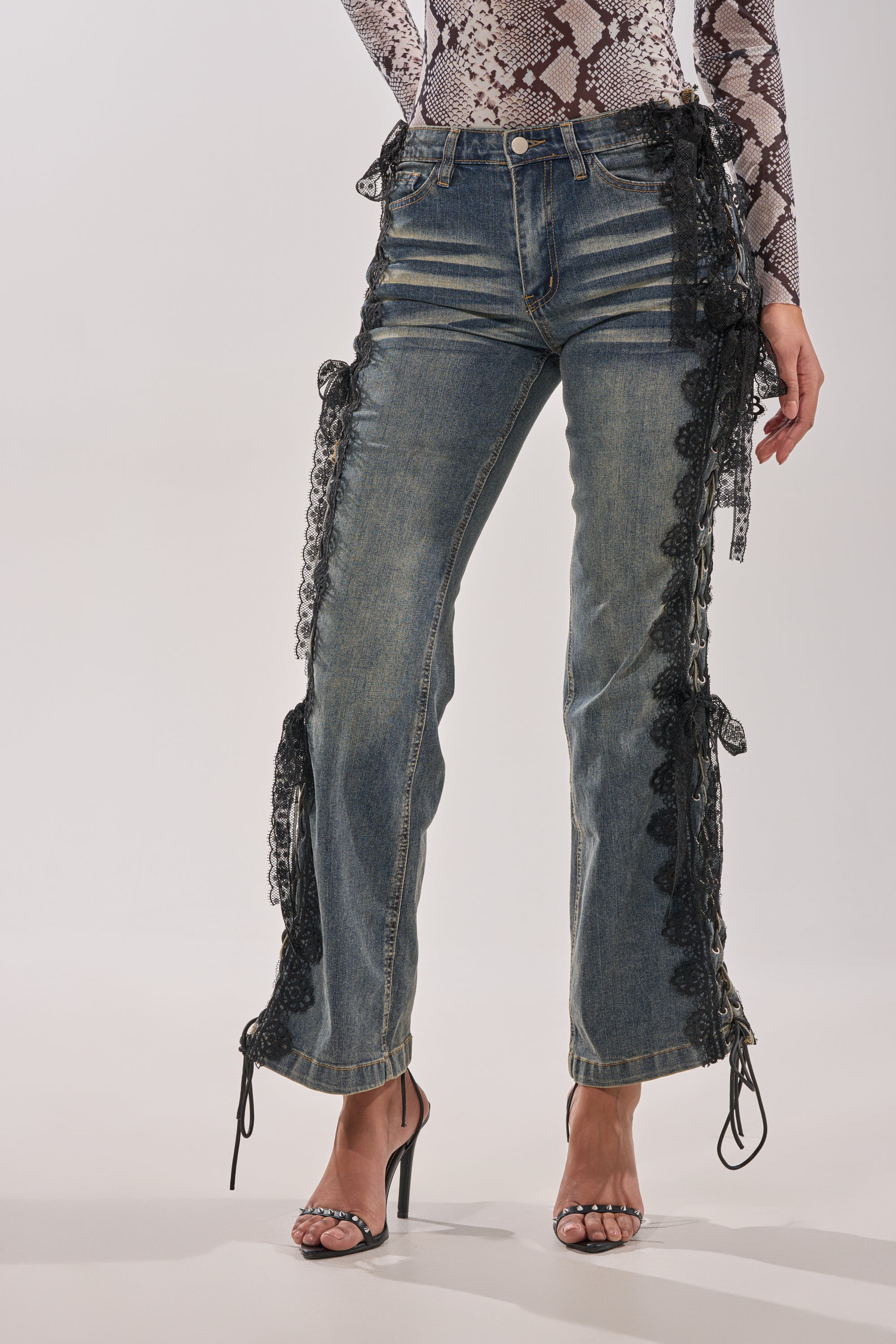 Wearing high-heeled sandals, ELIJAH FLARE LACE DENIM jeans with black lace-up sides, and a long-sleeve snakeskin top, a person stands against a plain background. 1