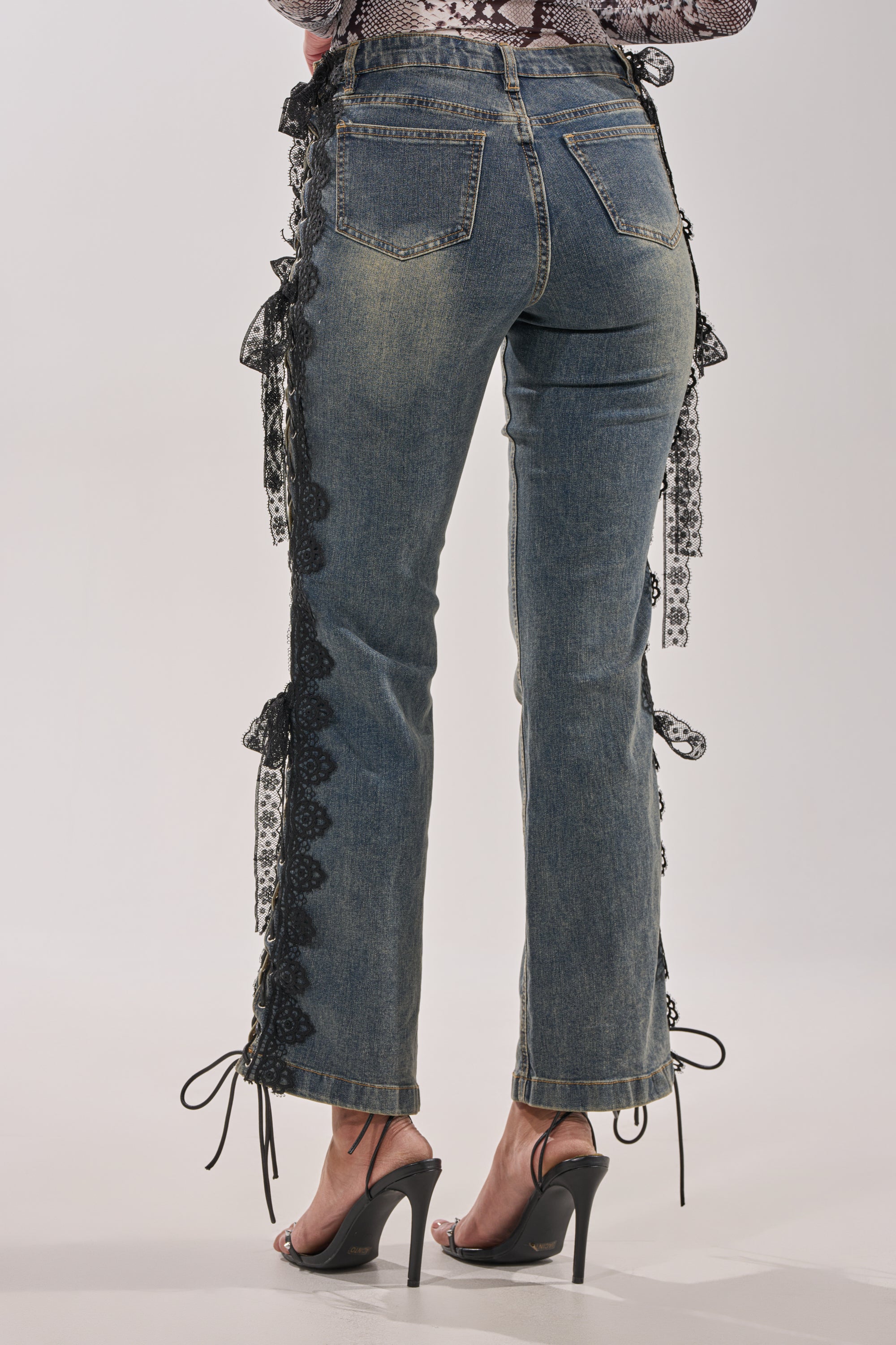 A person wears ELIJAH FLARE LACE DENIM with ribbon lace-up sides and black high-heeled sandals. The back view, waist down, highlights the trendy streetwear vibe of these stylish jeans. 3