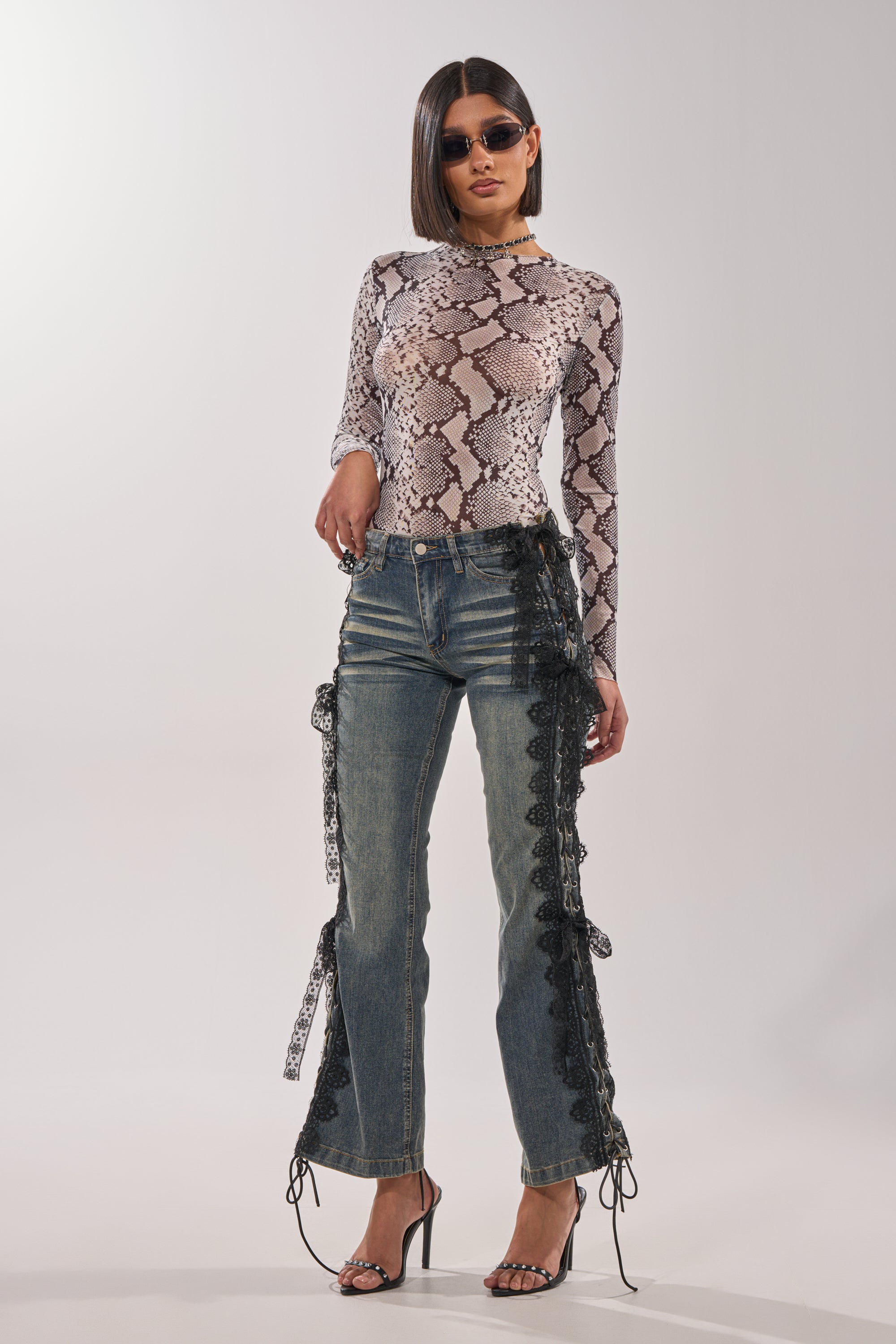 A woman wearing sunglasses, a snakeskin-print top, black heels, and ELIJAH FLARE LACE DENIM jeans stands against a plain light background. 4