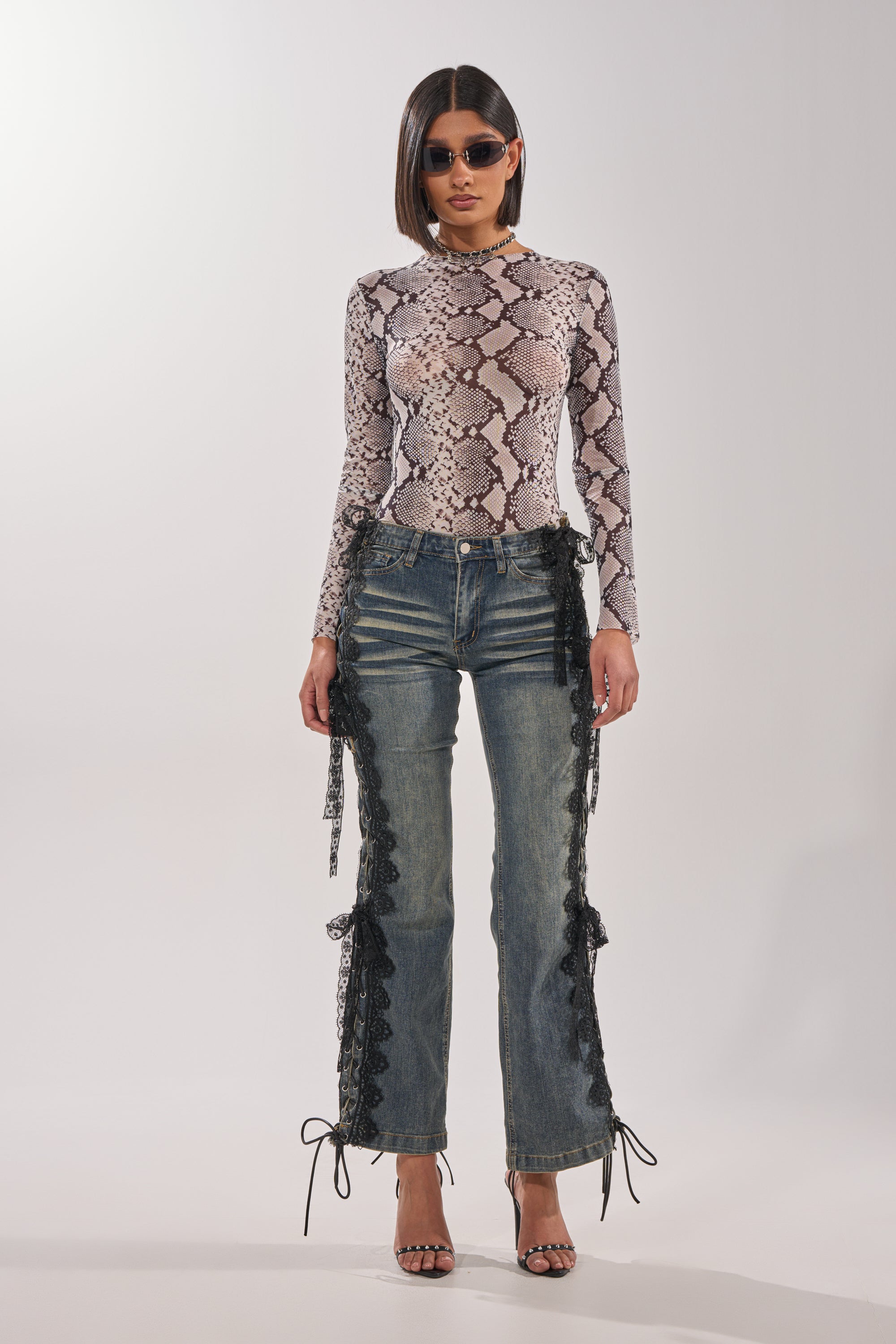 A woman wearing sunglasses, a long-sleeve snakeskin-print top, black strappy heels, and the ELIJAH FLARE LACE DENIM stands against a plain white background. 5
