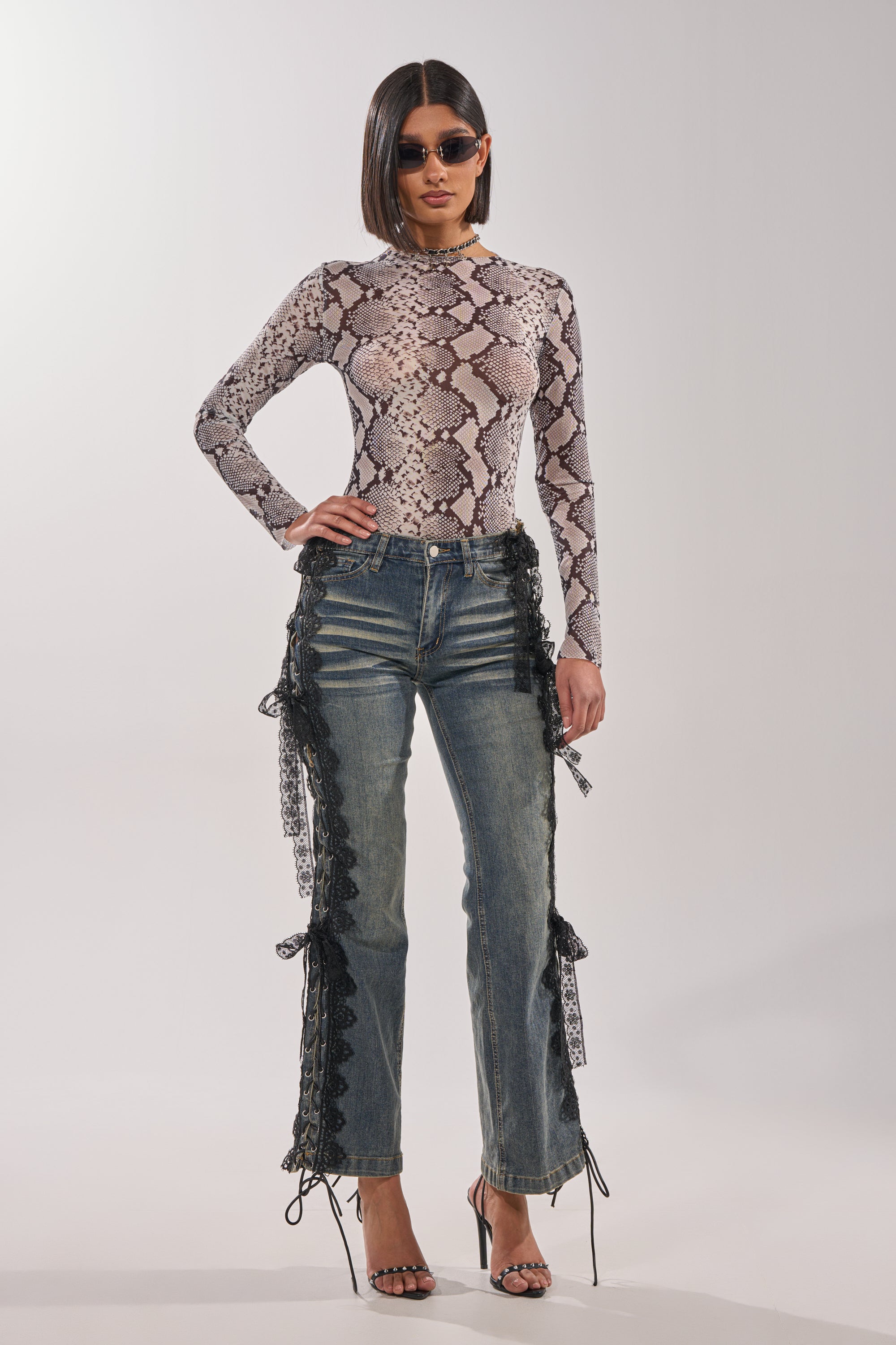 A woman wearing sunglasses, a snakeskin print top, black heels, and the ELIJAH FLARE LACE DENIM stands facing the camera against a light background. 7