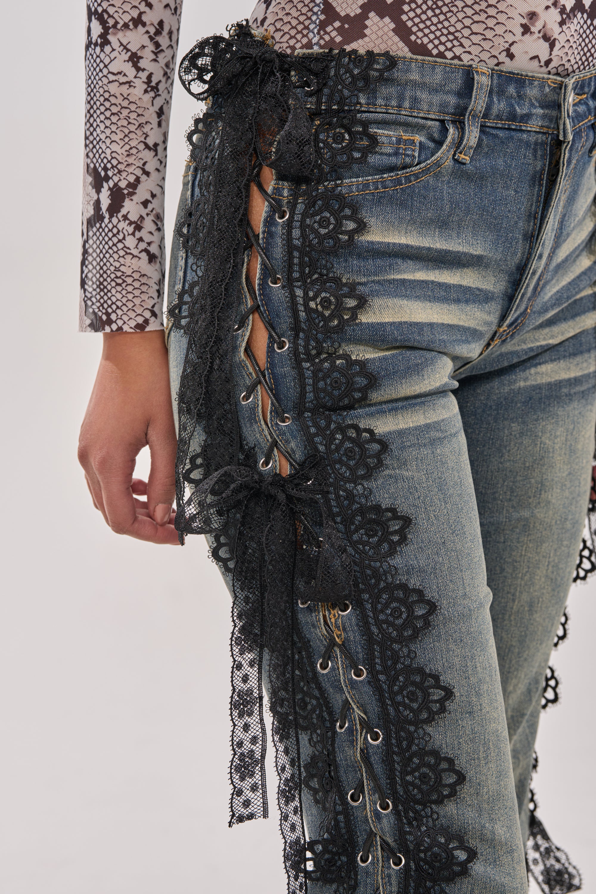 A close-up of someone wearing the ELIJAH FLARE LACE DENIM, featuring black ribbons laced through metal eyelets, paired with a gray and white snake print top for a bold, modern streetwear look. 8