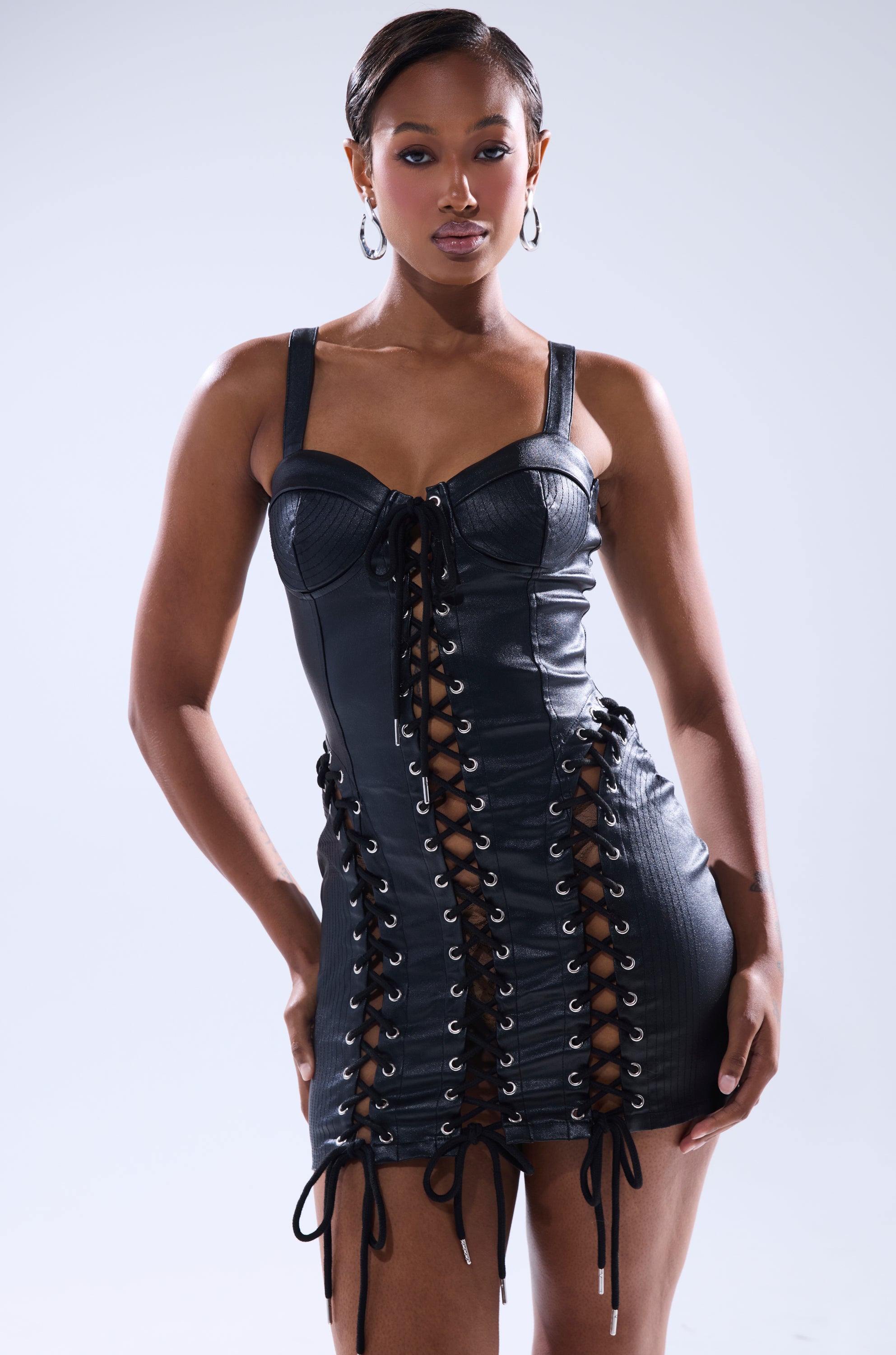 A woman stands confidently in the INTO THE NIGHT PU MINI DRESS—a black faux leather bodycon mini with grommet details—paired with hoop earrings, against a plain light background.