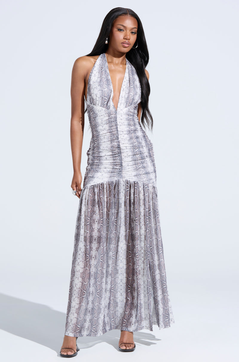 A woman with long, straight hair and heeled sandals poses against a white background in the BLAIR SNAKEPRINT MAXI DRESS, a sleeveless, floor-length halter dress with a plunging neckline and subtle snakeskin pattern.