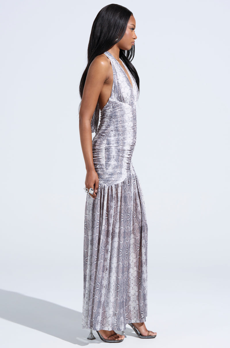 A woman poses in profile against a plain light background, wearing the BLAIR SNAKEPRINT MAXI DRESS— a sleeveless halter-style maxi with a deep V-neckline—paired with silver high-heeled sandals.