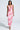 A woman stands confidently against a plain light background, wearing the VIOLETS FOR ROSES PRINTED MAXI DRESS in pink and white with a sleeveless design, straight hair, and neutral heels.