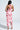 A woman with long straight hair models the VIOLETS FOR ROSES PRINTED MAXI DRESS, featuring a halter-neck, ruched design, and back slit. She stands with her back to the camera in strappy high heels.