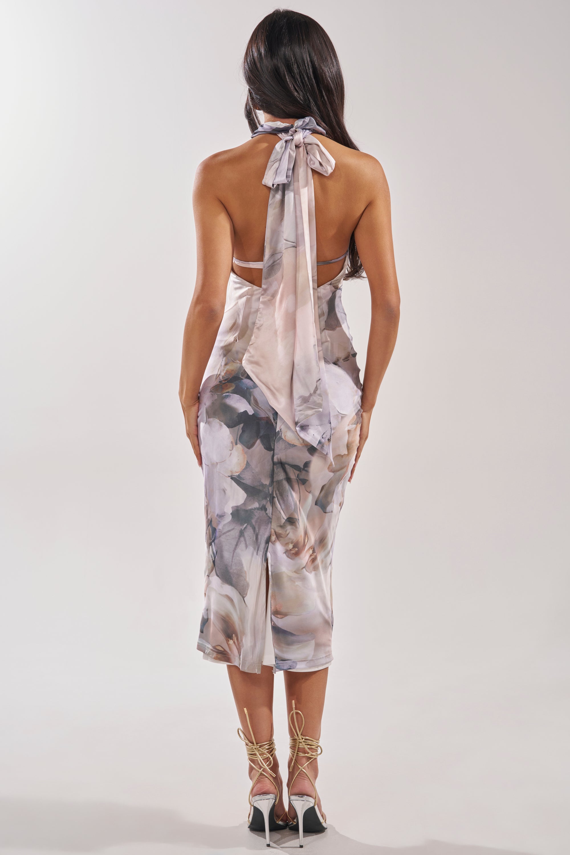 A woman with long dark hair is seen from the back wearing the SOFT GIRL ERA FLORAL MAXI DRESS, a halter neck, backless dress with a neck tie. She pairs it with lace-up high-heeled sandals against a plain, light background. 4