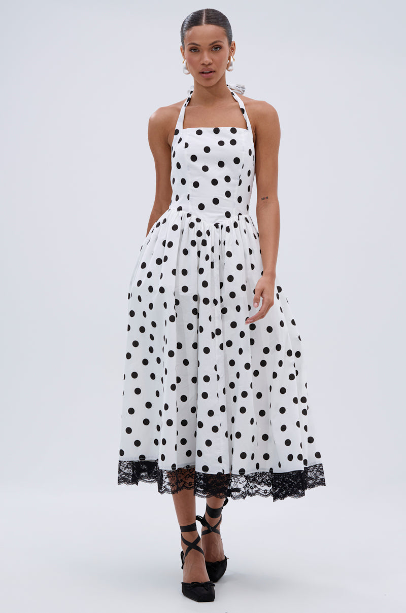 A woman wearing the DOLLY POLKA DOT MIDI DRESS with a white halter-neck and black lace trim, paired with black heels, stands against a plain light background.