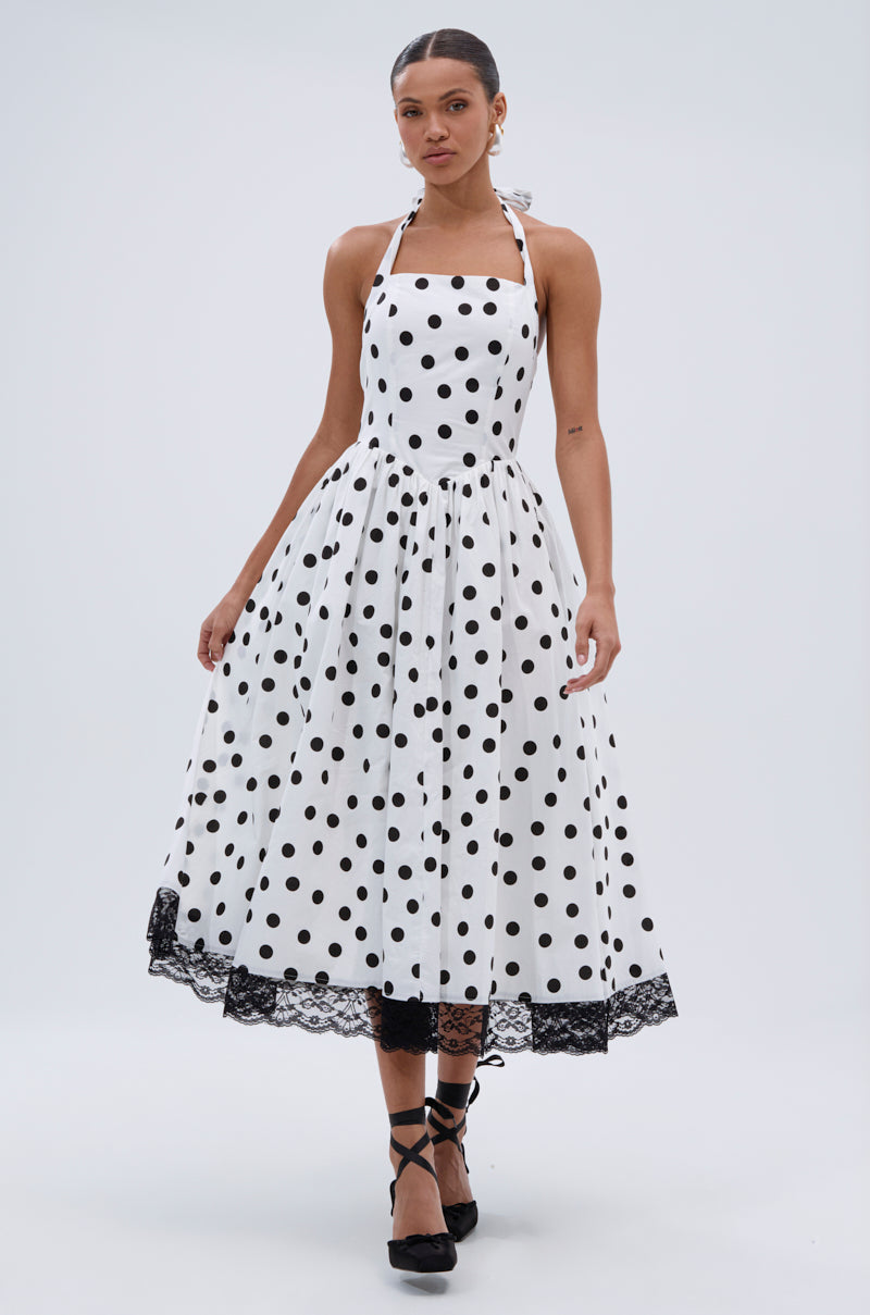 A woman models the DOLLY POLKA DOT MIDI DRESS—a sleeveless, white A-line halter dress with black polka dots, fitted bodice, flared midi skirt, and black lace hem—standing against a plain light background.