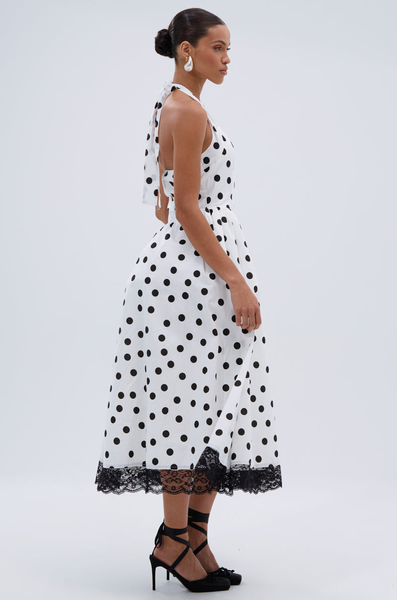 A woman in the DOLLY POLKA DOT MIDI DRESS—a white halter A-line midi dress with black lace trim—stands sideways. She pairs it with black heels, statement earrings, and a low bun for chic retro vibes.