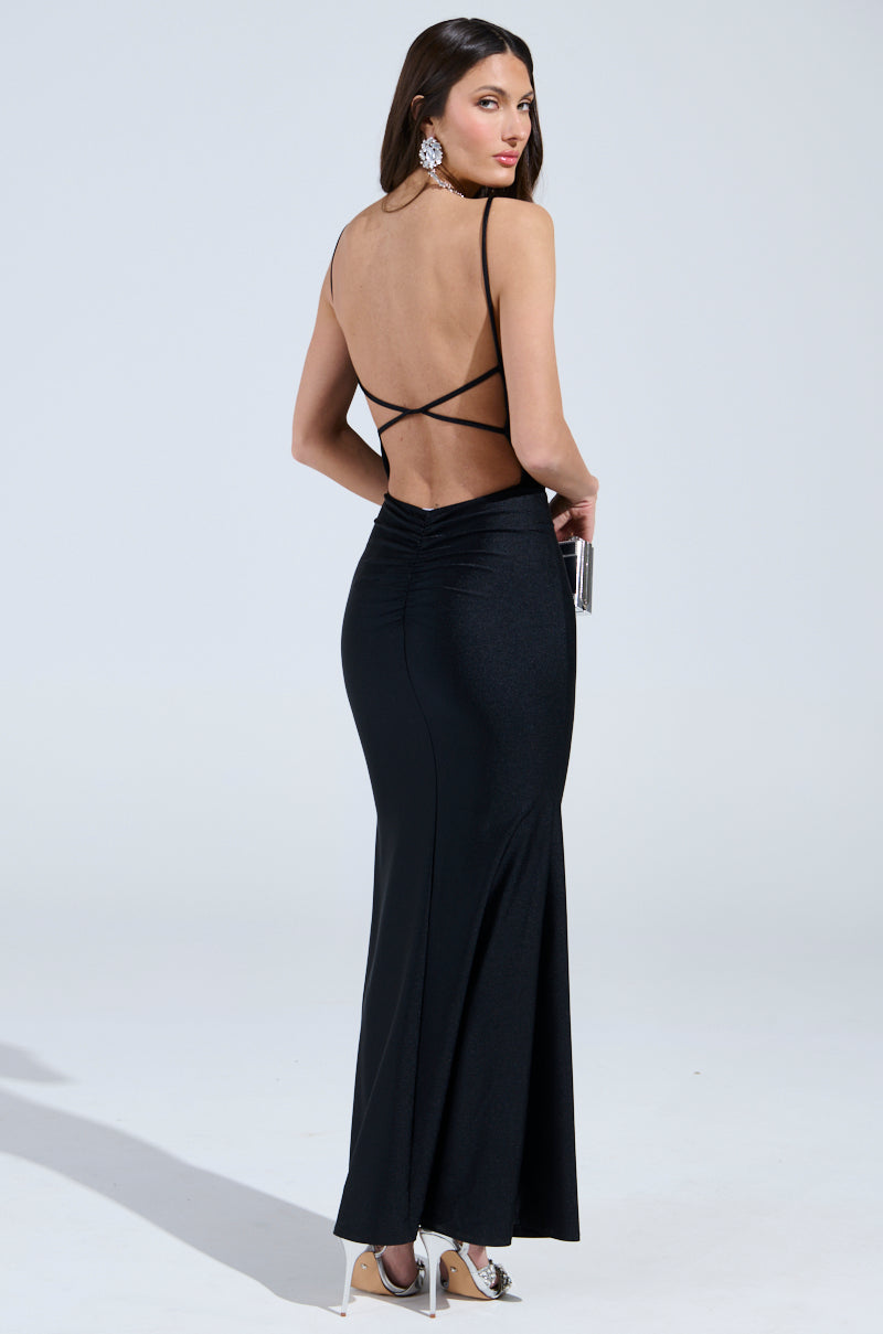 A woman gracefully wears the "Lady of the Night" cowl neck maxi dress in black, featuring a long, backless design with thin straps. She accessorizes with a silver clutch and large earrings, enhancing her elegant style against a white backdrop.