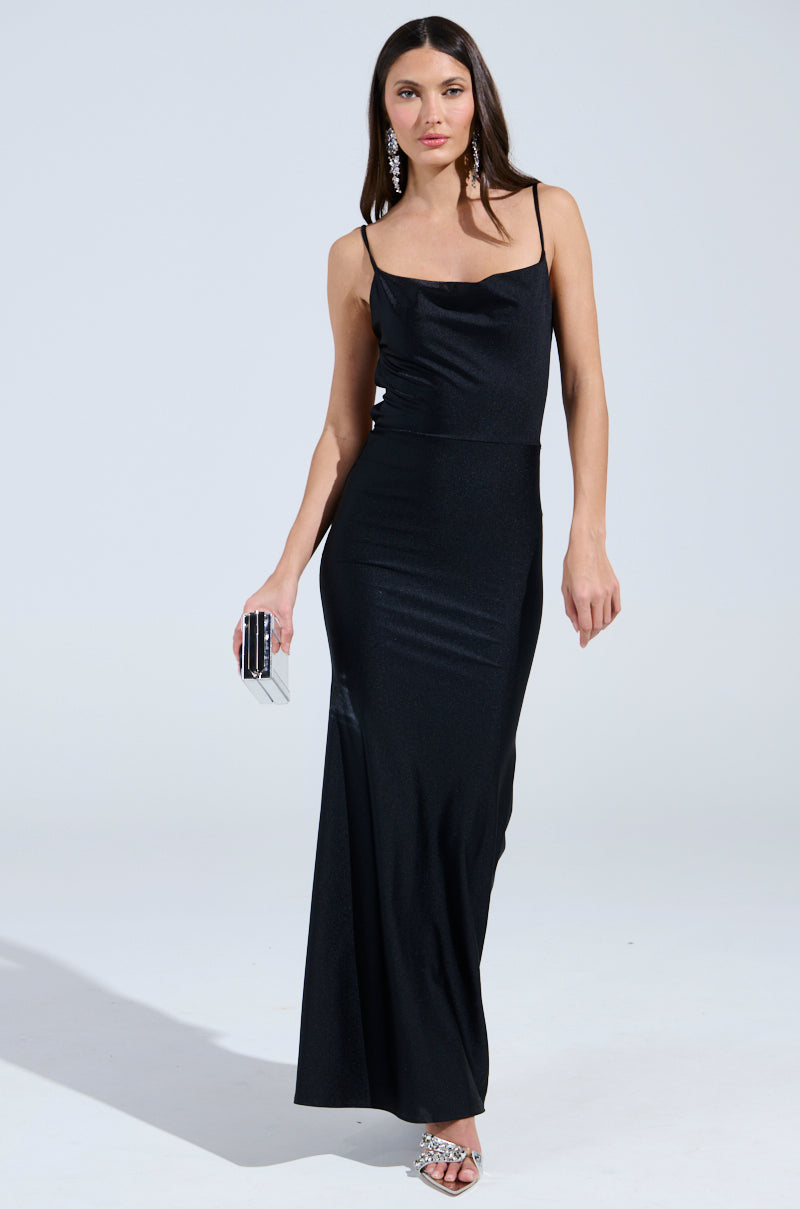 Dressed in the LADY OF THE NIGHT COWL NECK MAXI DRESS IN BLACK, a woman accessorizes with a small clutch, strappy high heels, and large earrings. The fitted silhouette and thin straps add elegance as she poses against a neutral background.