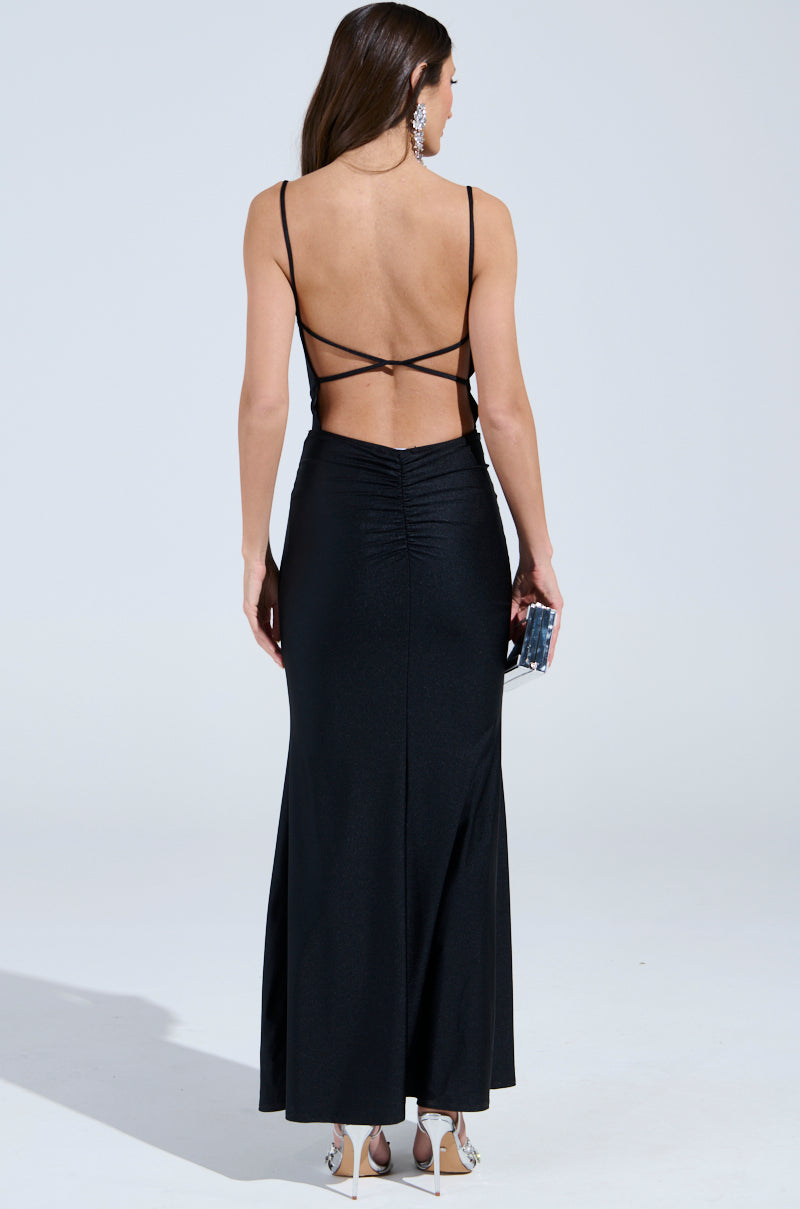 A woman poses with her back to the camera in the LADY OF THE NIGHT COWL NECK MAXI DRESS IN BLACK, featuring thin straps and a sleek design. Her loose hair cascades elegantly as she holds a small clutch, wearing silver high heels and large dangling earrings.