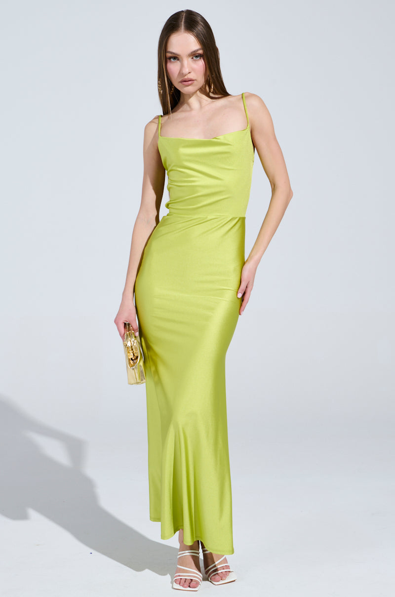 A woman models the Lady of the Night cowl neck maxi dress in olive, elegantly holding a small gold clutch and wearing strappy white heels. Her straight long hair is styled against a plain light gray background, casting a soft shadow beside her.