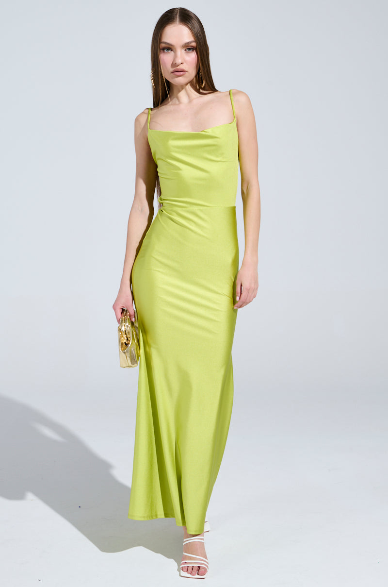 A woman stands on a plain white background in the LADY OF THE NIGHT cowl neck maxi dress in olive, holding a small gold purse. Her long straight hair flows down her back while wearing white high-heeled sandals.