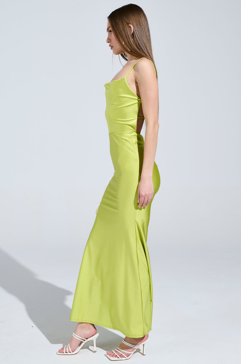 A person with long hair models the LADY OF THE NIGHT COWL NECK MAXI DRESS in olive, paired with white strappy high heels, standing elegantly in profile against a plain, light background.