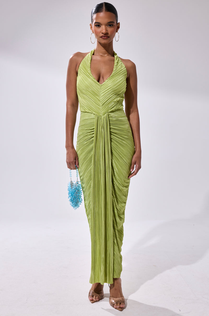 A woman stands against a plain background in the SARAH SLINKY DRESS IN GREEN, a light green pleated maxi dress with a draped halter-neck front. She accessorizes with hoop earrings, clear high-heeled sandals, and a small blue beaded handbag.