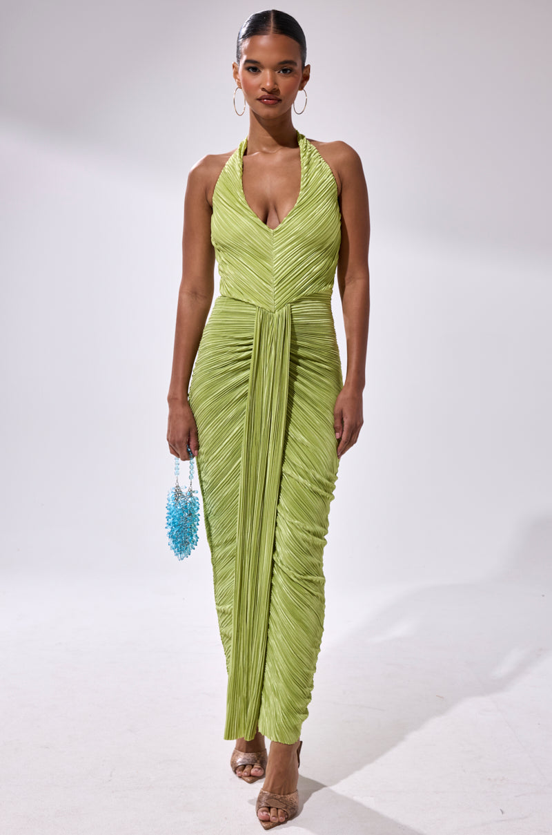 A woman in the SARAH SLINKY DRESS IN GREEN with clear high heels stands against a plain background, holding a small blue beaded handbag.