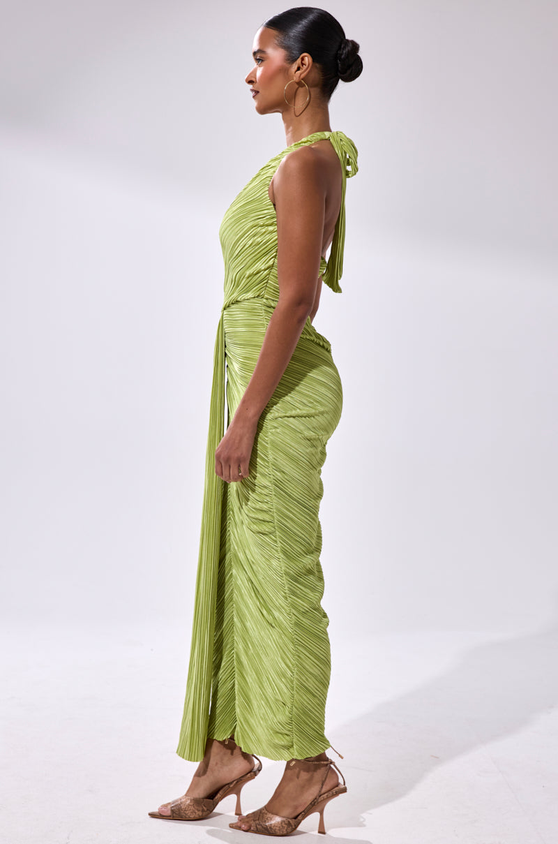 A woman stands in profile wearing the SARAH SLINKY DRESS IN GREEN, a light green pleated gown with draped details, paired with brown strappy heels. Her hair is styled in a sleek bun as she poses against a plain white background.
