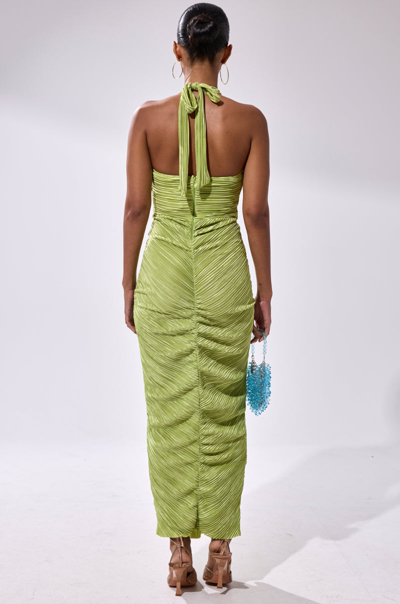 A woman faces away in the SARAH SLINKY DRESS IN GREEN with tan heels, a blue beaded handbag, hair in a bun, and large hoop earrings against a plain light background.