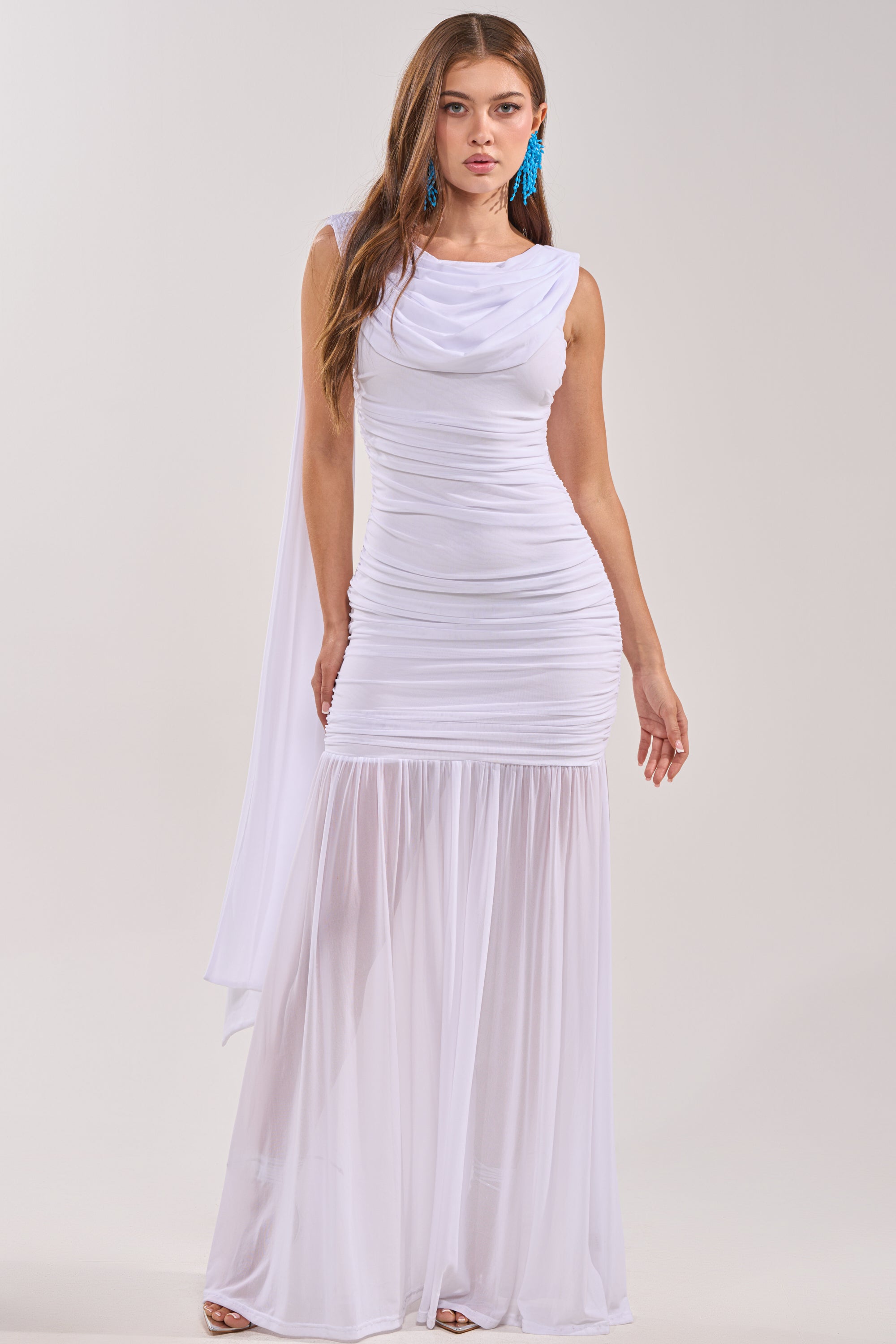 A woman poses against a plain background in the TRENDSETTER ENERGY Layered Maxi Dress in white, featuring sheer mesh from the knees down and gathered bodice detailing—styled with blue statement earrings for an elegant, bridal-ready look.