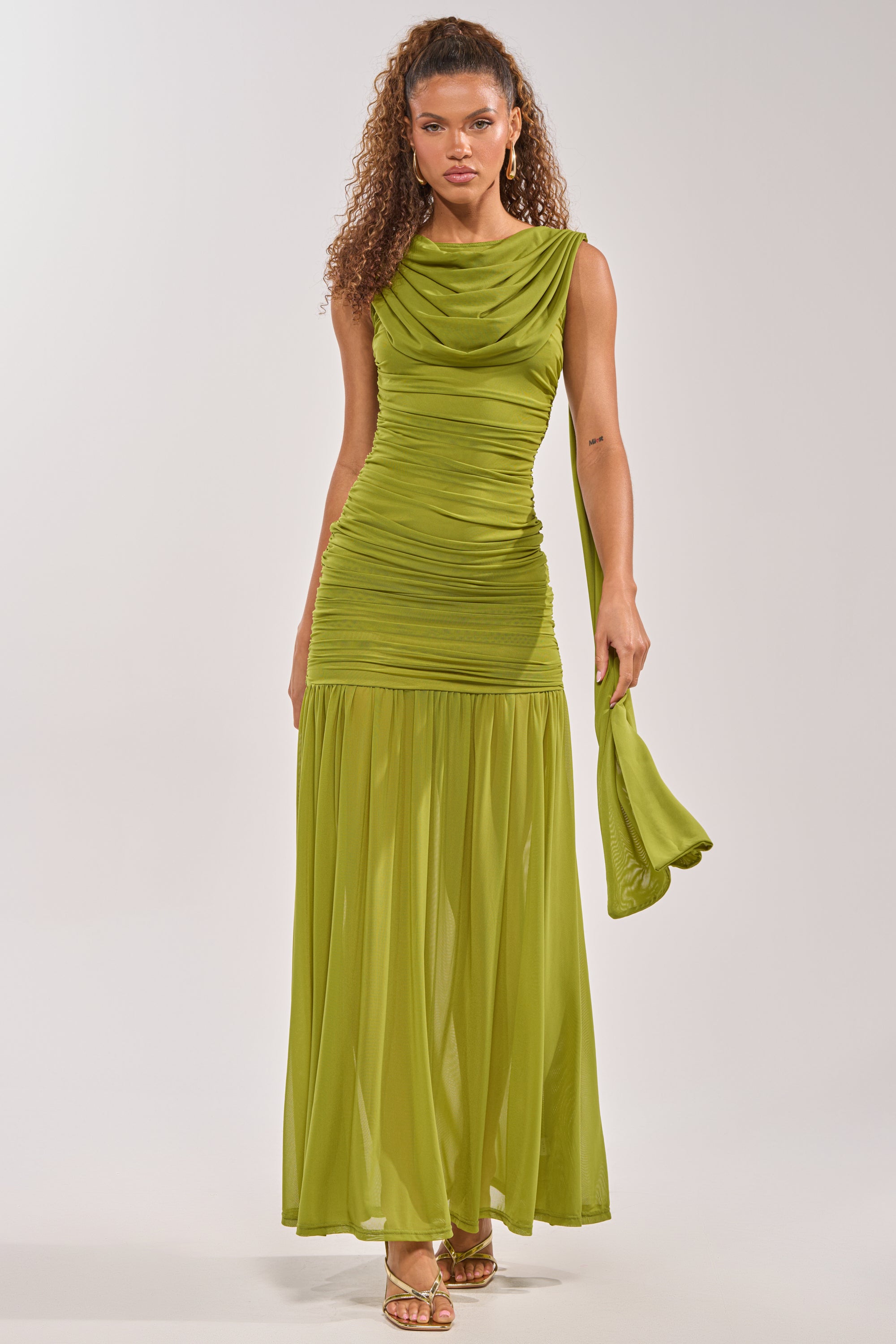 A woman with curly hair wears the TRENDSETTER ENERGY Layered Maxi Dress in green—sleeveless with ruched details, a sheer skirt, and a matching scarf—paired with gold heels against a plain background. 0