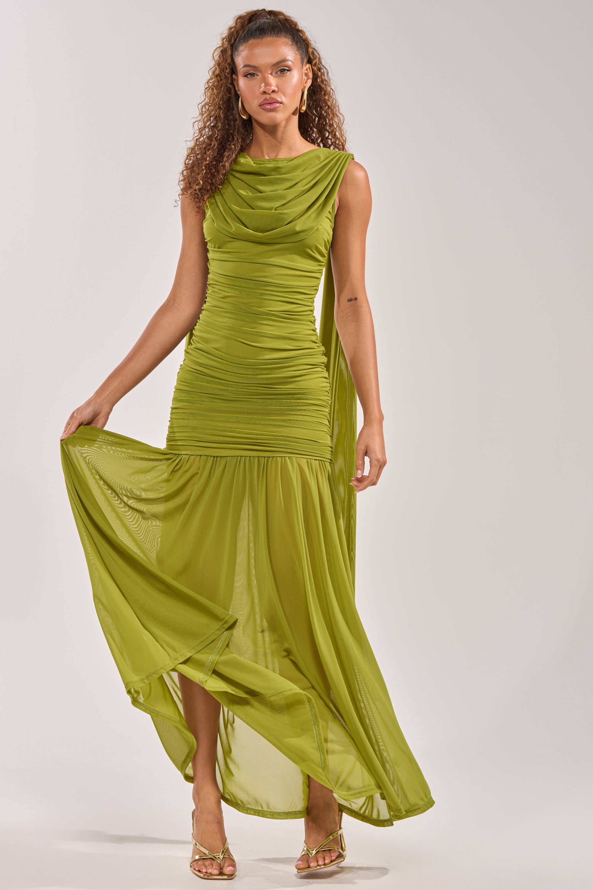 A woman with curly hair and gold earrings holds out one side of the TRENDSETTER ENERGY LAYERED MAXI DRESS IN GREEN, a sleeveless ruched wedding guest dress with a flowing, sheer skirt. She pairs it with gold high-heeled sandals. 1