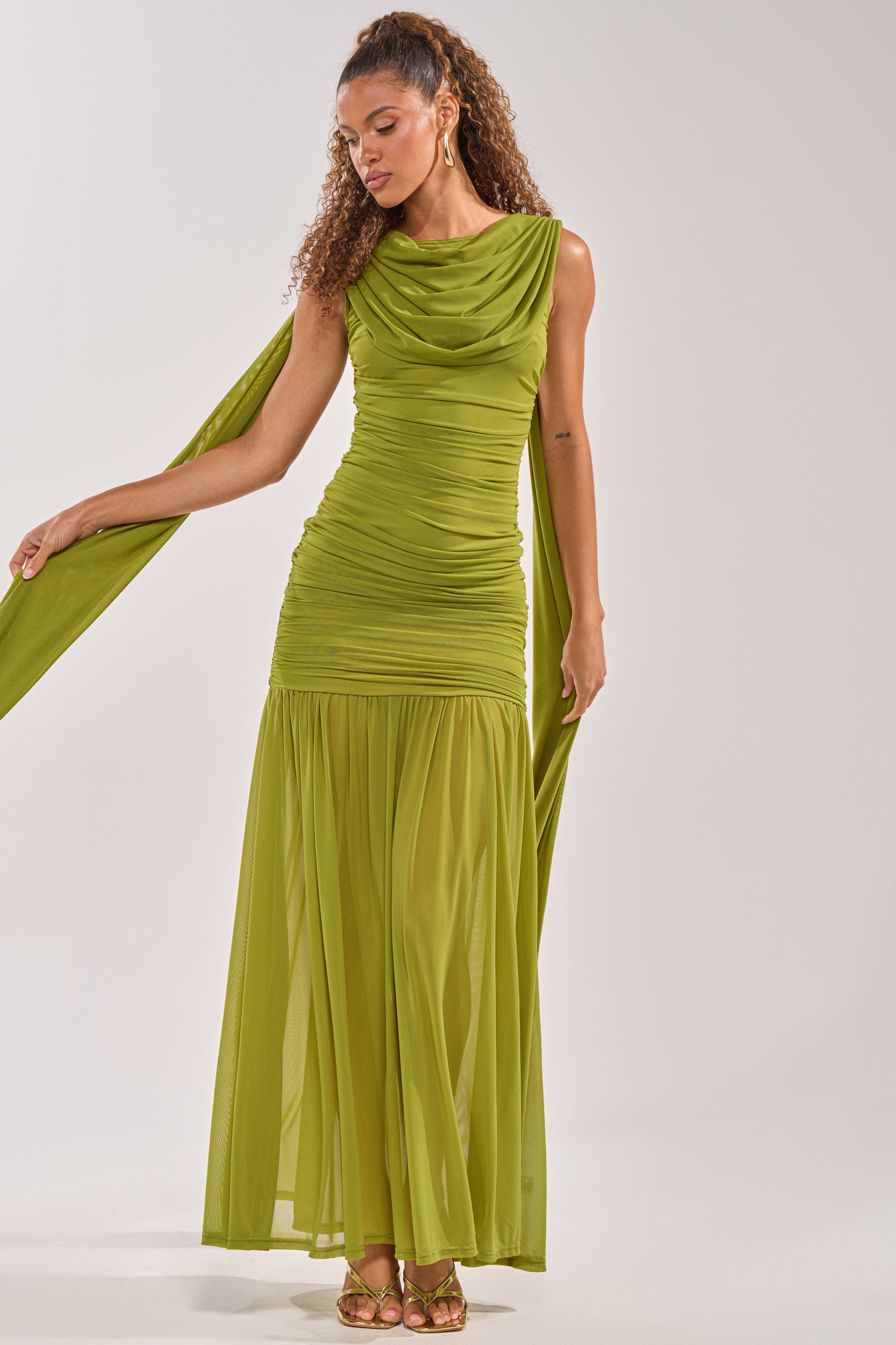 A woman in the TRENDSETTER ENERGY Layered Maxi Dress in green, a sleeveless, ruched, floor-length formal dress with a draped scarf, stands on a plain light background, looking down and holding the scarf with both hands. 2