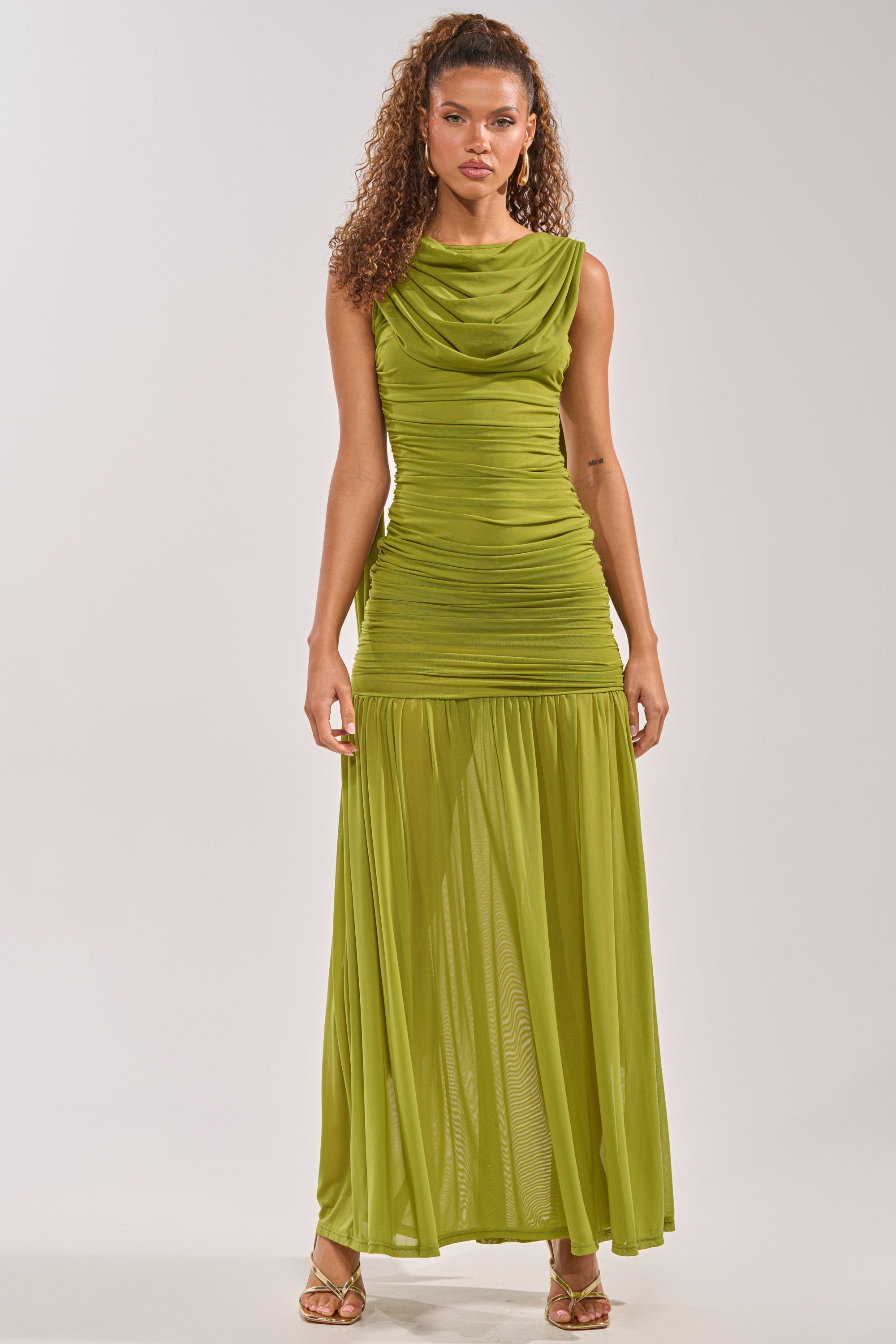 Wearing the TRENDSETTER ENERGY LAYERED MAXI DRESS IN GREEN, a woman stands facing forward. This sleeveless, floor-length dress with a draped neckline and ruched bodice is perfect for weddings, beautifully styled with curly hair and gold sandals. 3