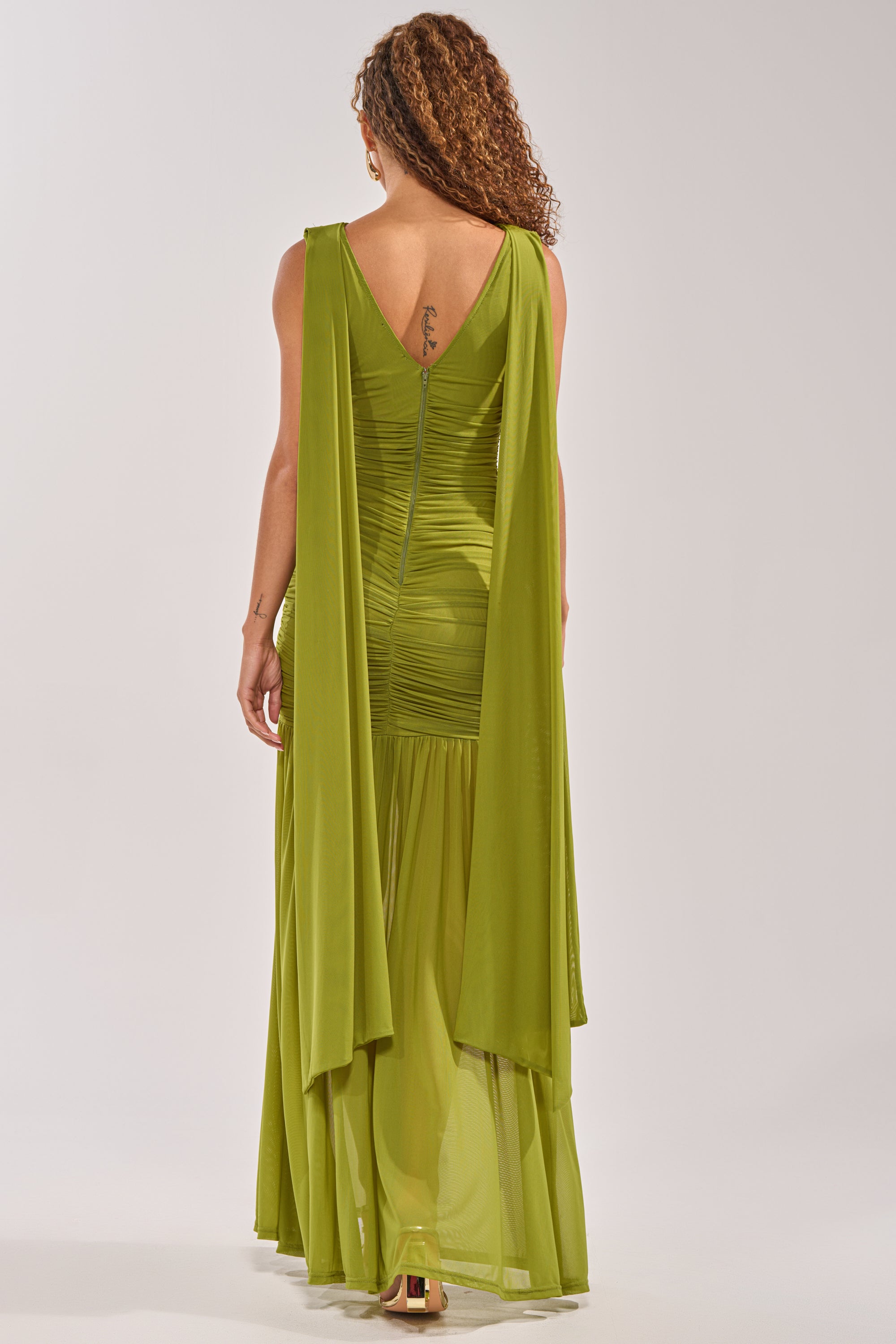 A woman with curly hair shows a tattoo on her upper back while wearing the TRENDSETTER ENERGY LAYERED MAXI DRESS IN GREEN—a sleeveless, floor-length mesh mermaid dress ideal for weddings or any formal event. 5