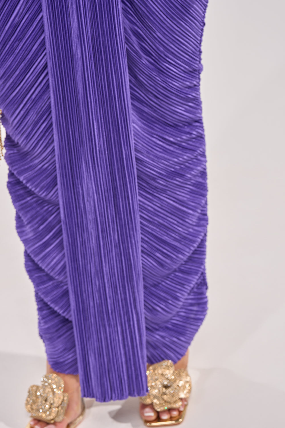 A person wearing the TILL SUNRISE MAXI DRESS IN DEEP PURPLE, featuring pleats and a halter neckline, with gold embellished open-toe sandals, stands on a light-colored surface. 4