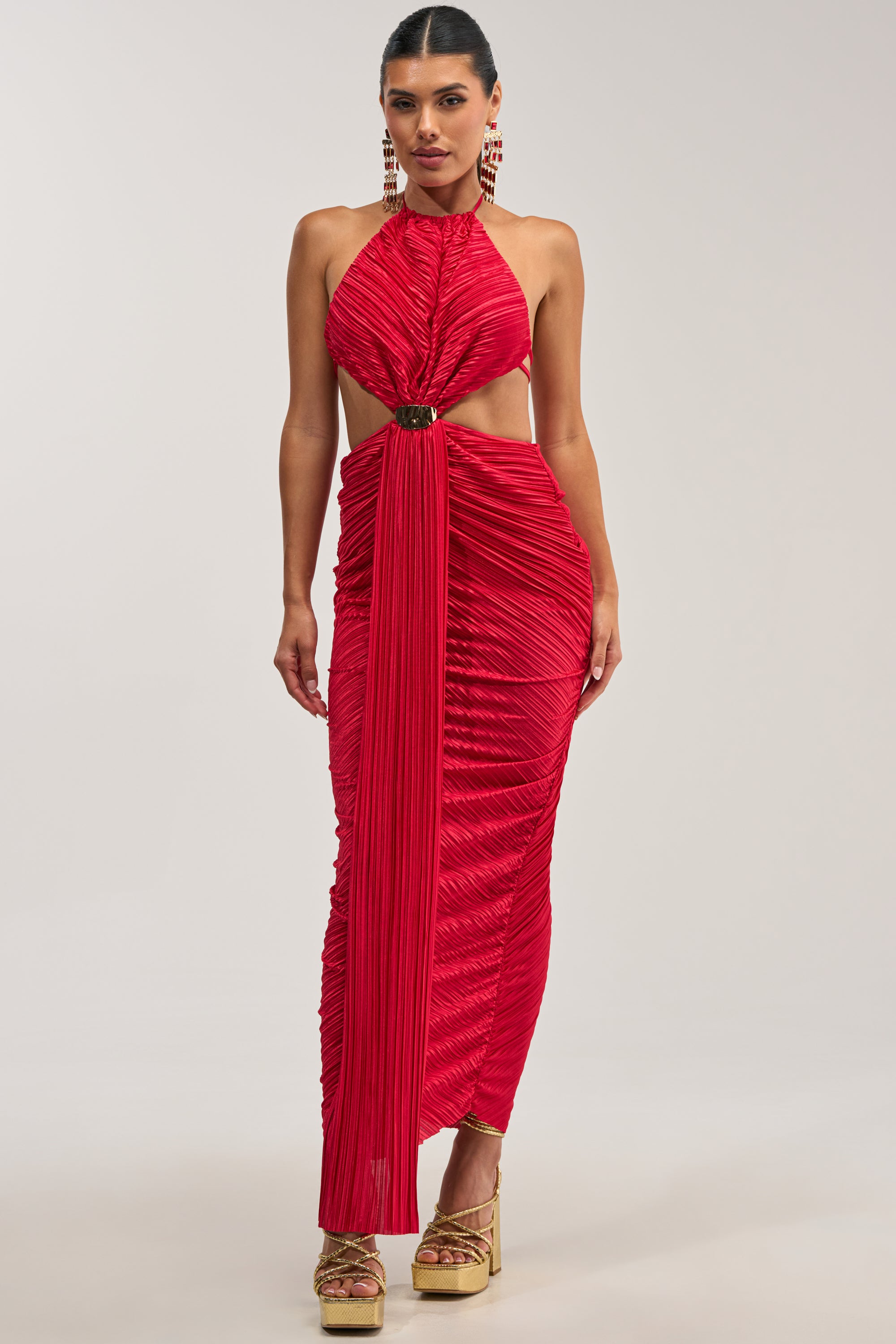 Dressed in the TOO CUTE TO ARGUE SLINKY MAXI DRESS—a sleeveless, ruched red satin halter maxi featuring waist cutouts and gold buckle detailing—this woman stands in platform heels against a plain light background, making a striking statement as a wedding guest or at an elegant evening event.