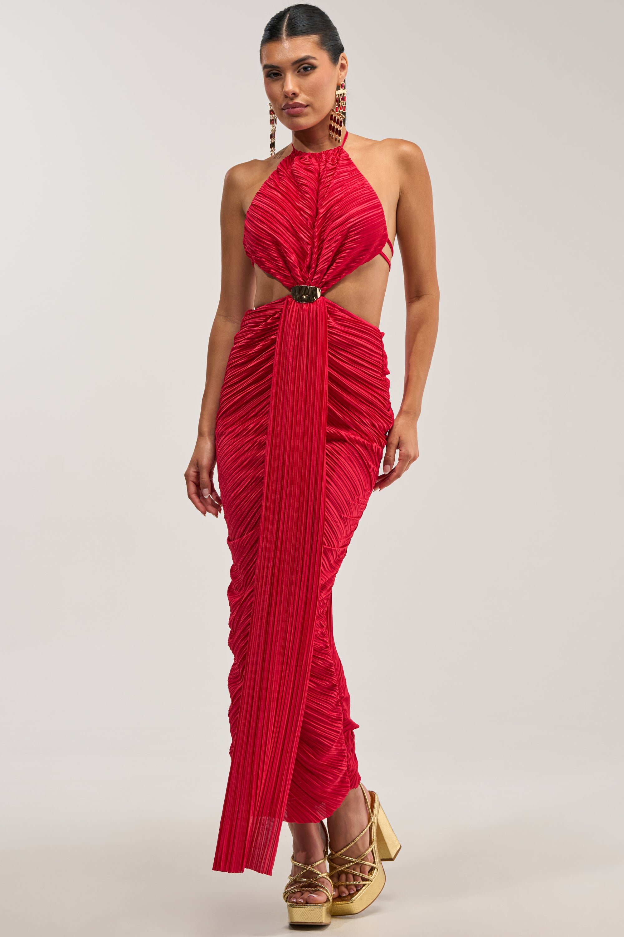 Showcasing the TOO CUTE TO ARGUE SLINKY MAXI DRESS—a sleeveless, ruched red satin halter maxi dress featuring a front cutout and long center panel. Styled with gold earrings and gold platform heels against a plain background, this ensemble is an elegant option for evening events or as a wedding guest dress.