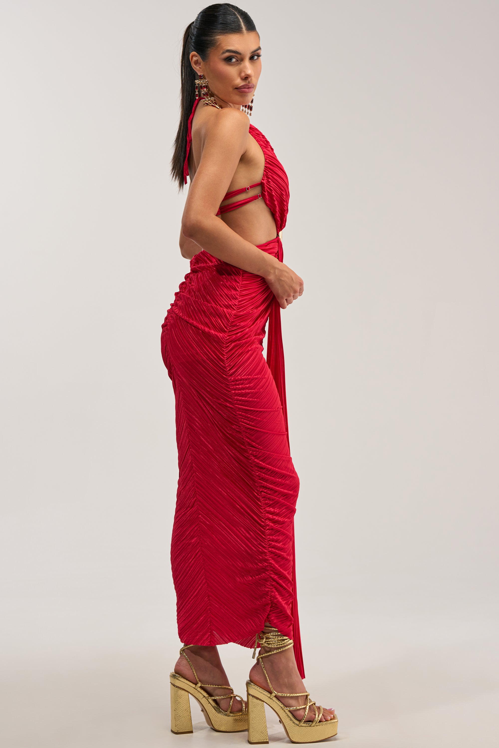 Dressed in the TOO CUTE TO ARGUE SLINKY MAXI DRESS—a vibrant red satin halter maxi featuring ruching and side cutouts—she pairs the look with gold platform heels and poses against a simple, light backdrop, glancing over her shoulder. This statement dress is perfect for an elegant evening out or to turn heads as a wedding guest.