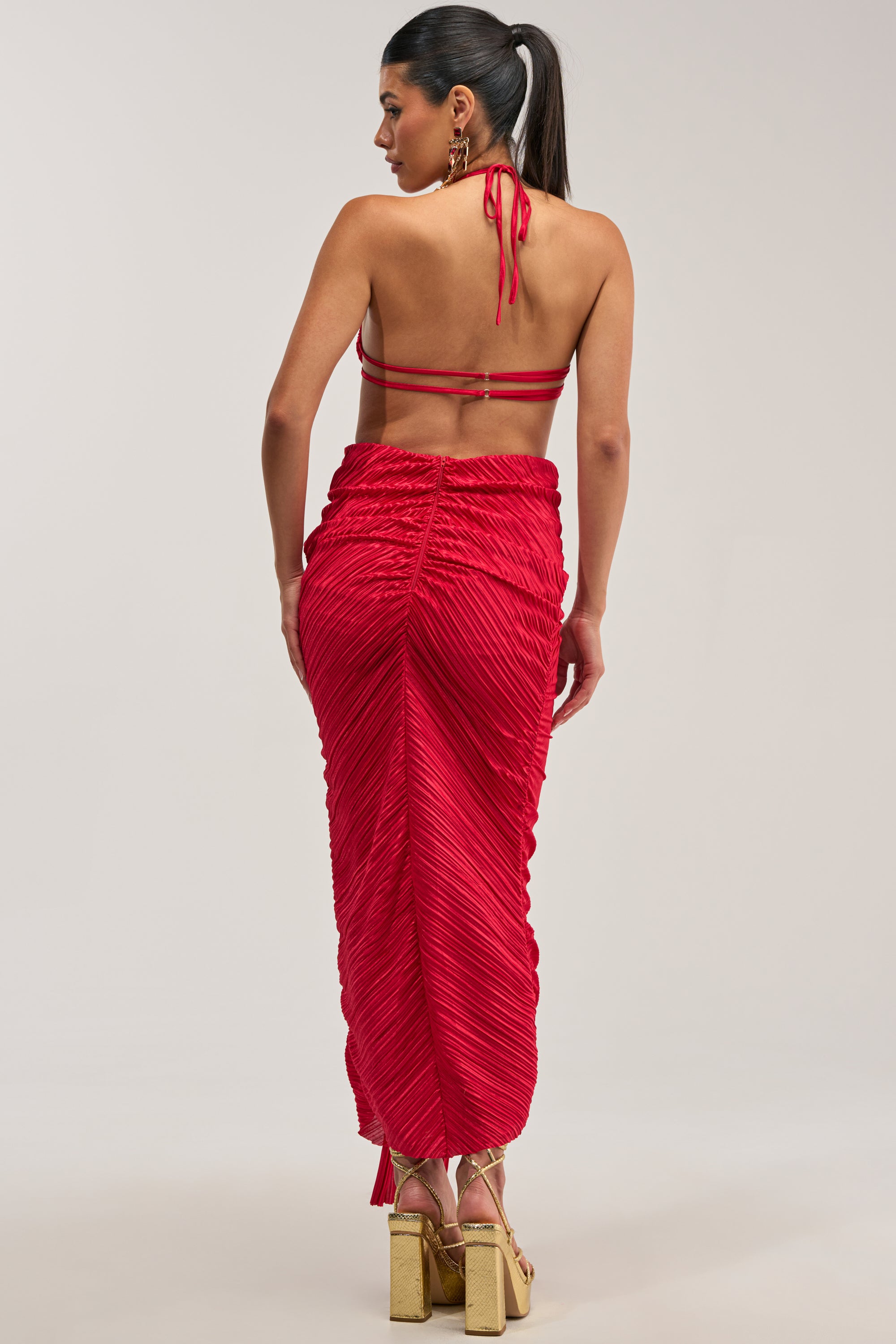 Wearing the TOO CUTE TO ARGUE SLINKY MAXI DRESS, a woman with a high ponytail is seen from behind in a dramatic red halter-neck maxi silhouette with a backless design and graceful pleats. She completes her evening-ready ensemble with gold strappy platform heels against a plain, light gray background—an effortlessly chic look ideal for an evening gown or as the perfect wedding guest dress.