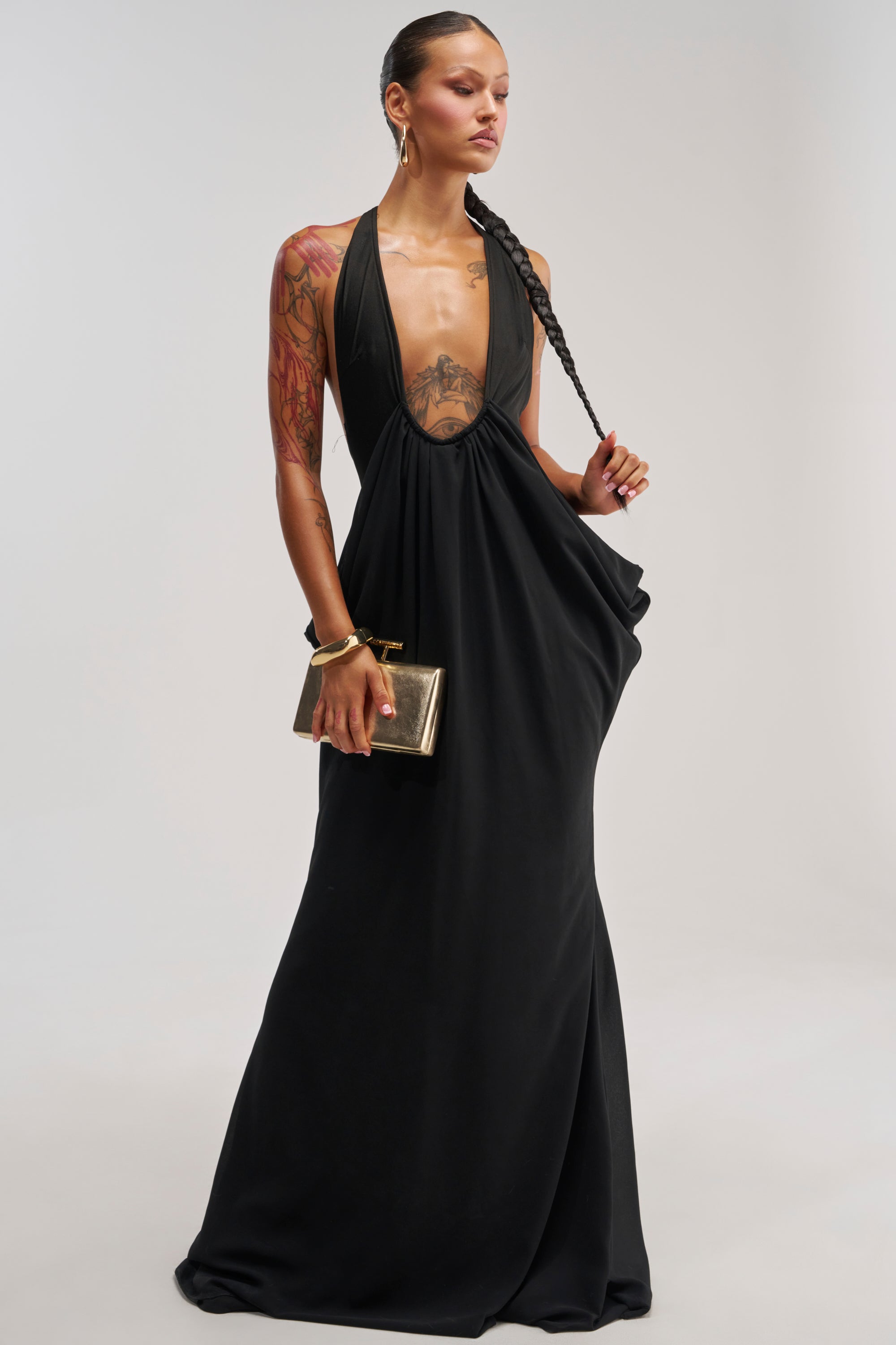 A woman wearing the BORN TO SHINE SWOOP MAXI DRESS IN BLACK, featuring a halter neckline, stands against a plain light background. She accessorizes with hoop earrings and a small gold clutch, her hair styled in braids and tattoos visible on her arm.