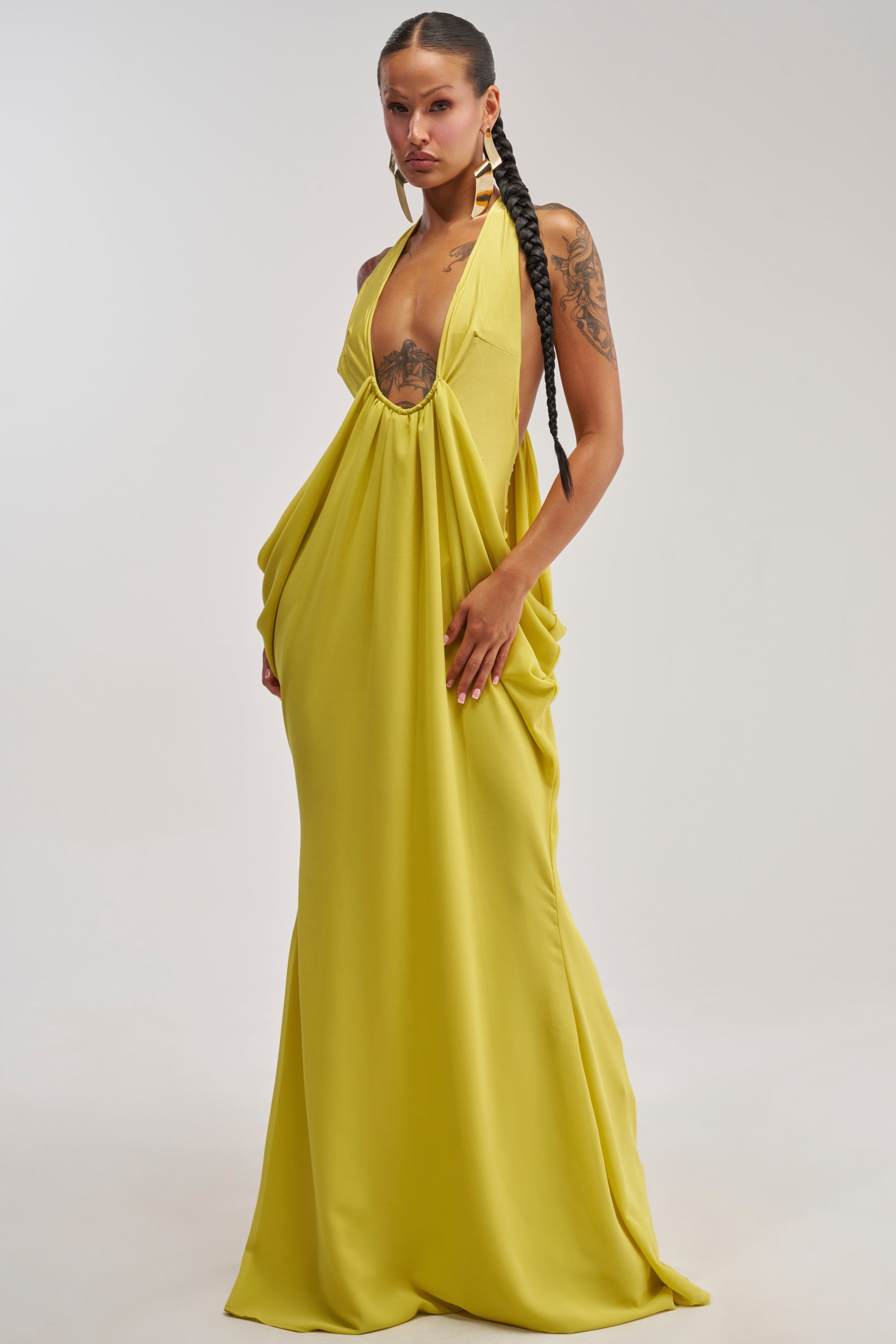 A woman poses in the BORN TO SHINE SWOOP MAXI DRESS, a long and flowing chartreuse gown featuring draped sides and a halter-neck design. She has tattoos on her arm and chest, accessorized with large earrings, set against a simple white background.