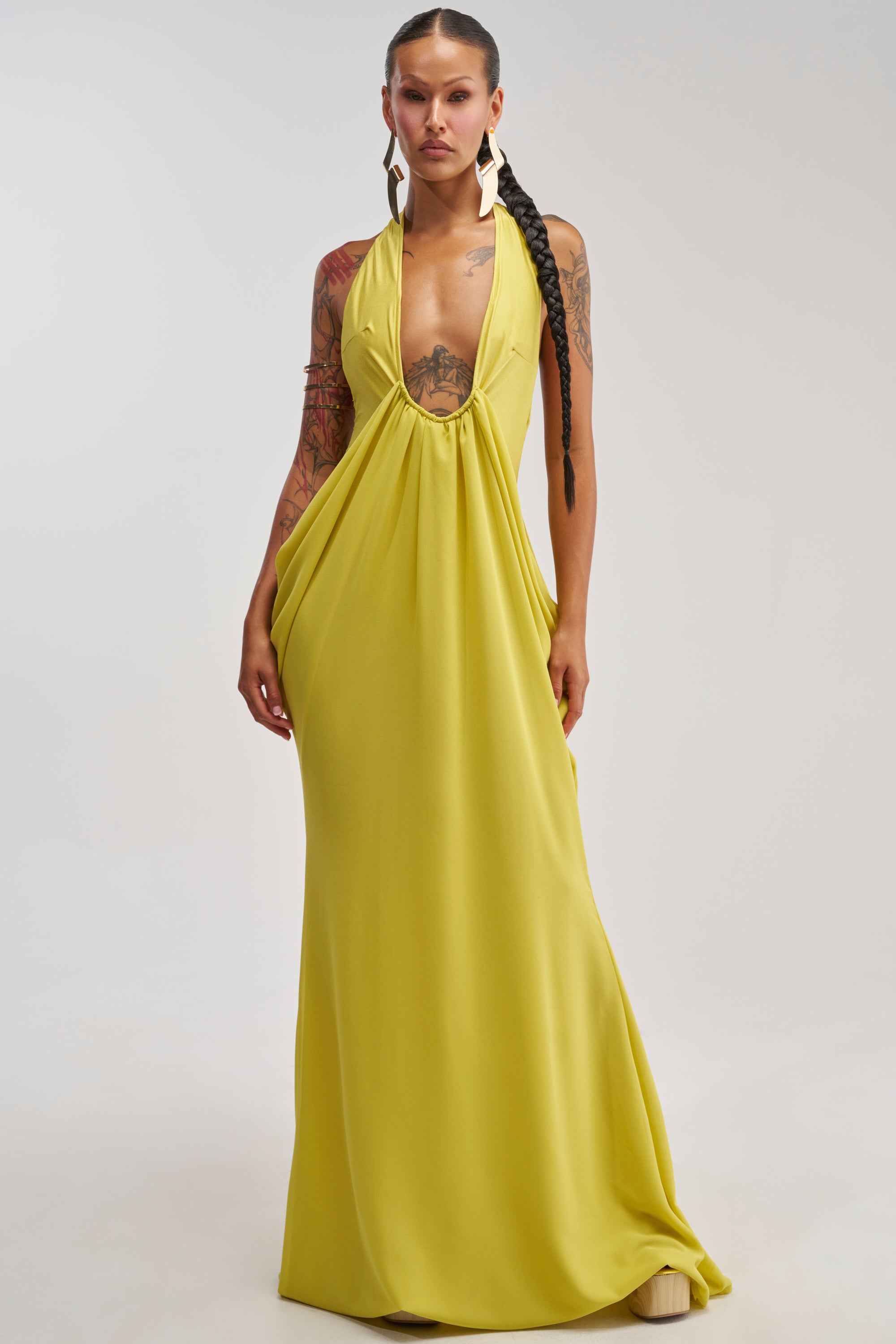 A person wearing the BORN TO SHINE SWOOP MAXI DRESS, a long, sleeveless chartreuse maxi dress featuring a deep plunging neckline, poses against a plain light background. They have long braided hair, visible tattoos, and accessorize with large white earrings.